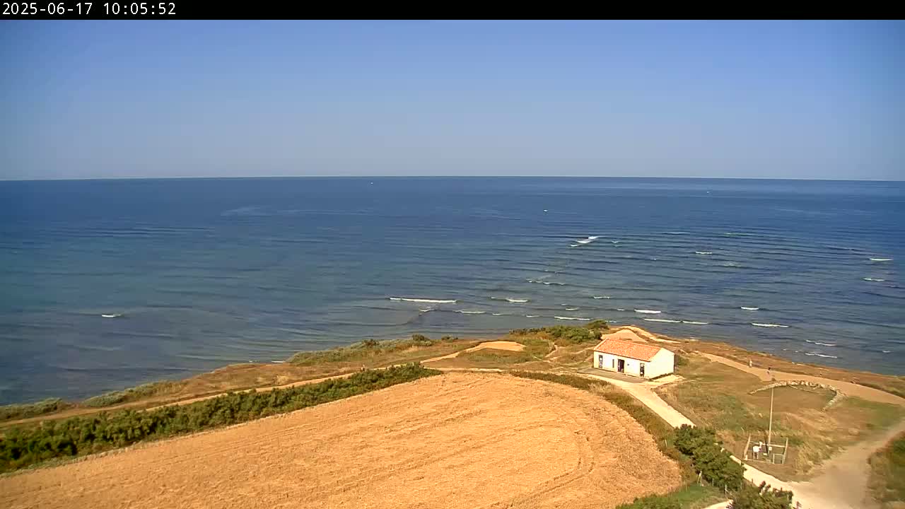A small white building sits on a grassy hill overlooking a calm ocean under a clear blue sky.