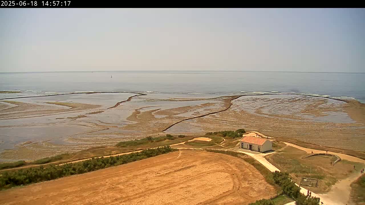A high-angle view shows a small building near a calm ocean under a clear sky, with low-lying tidal pools and a brown field visible in the foreground.