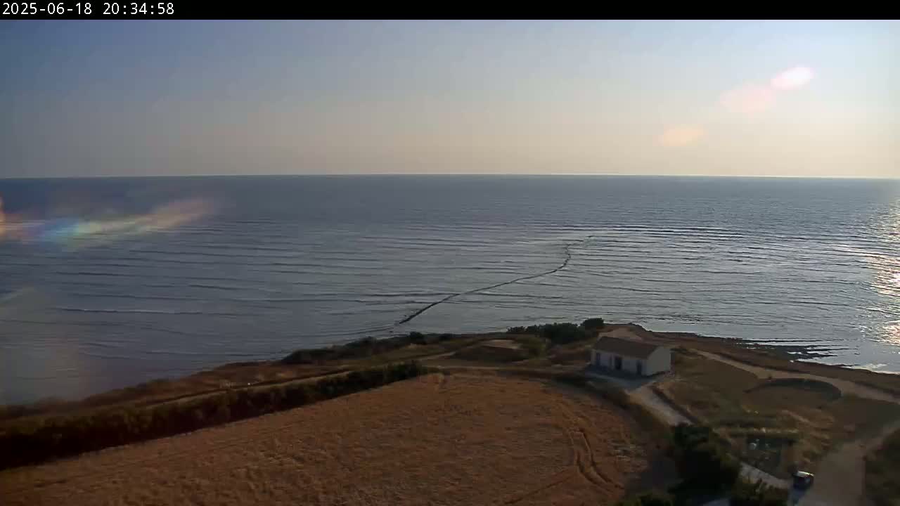 A high-angle view shows a calm sea under a clear sky, with a small house situated on a coastal cliff overlooking a brown field.