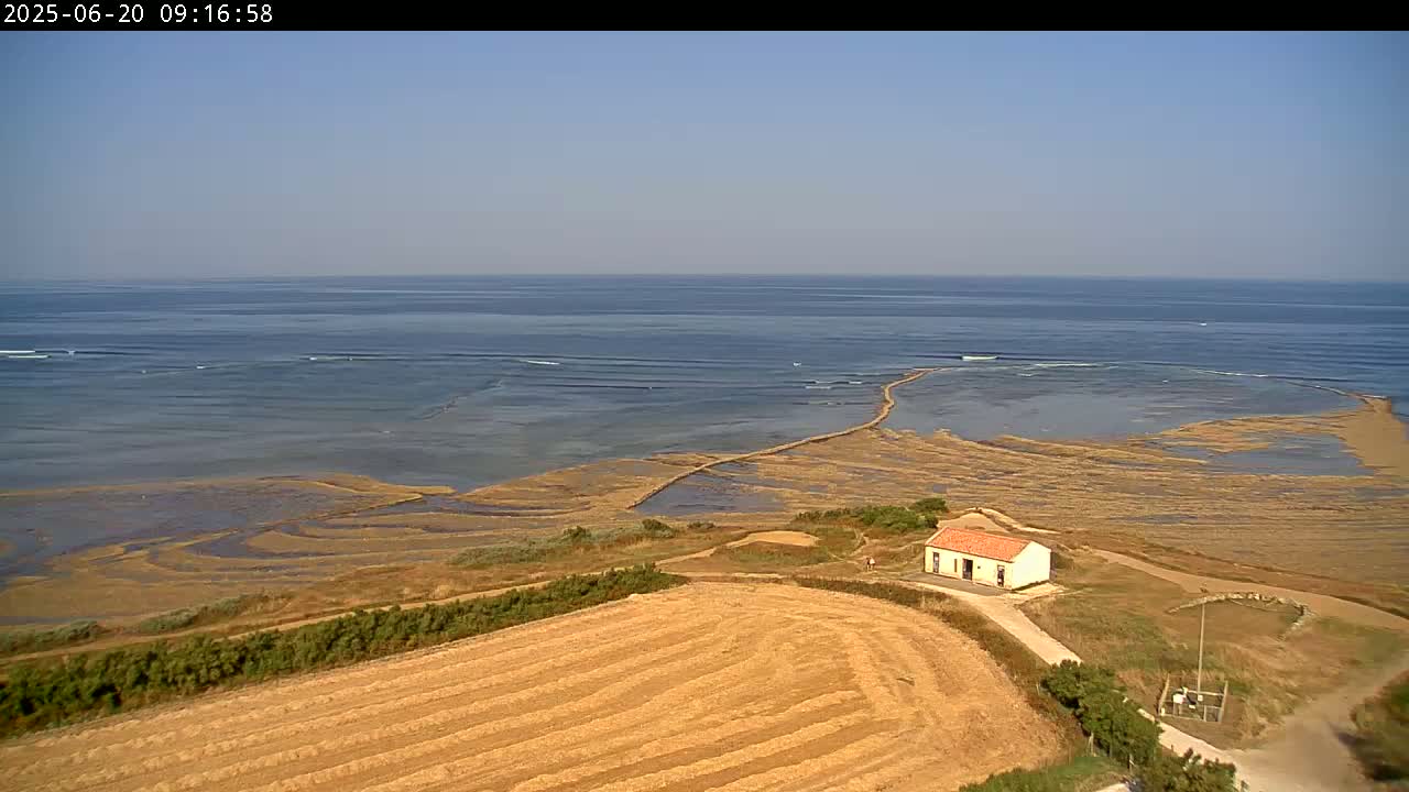A small, light-colored building sits on a grassy hillside overlooking a coastal area with a long, shallow, sandy expanse leading to a calm ocean under a clear sky.