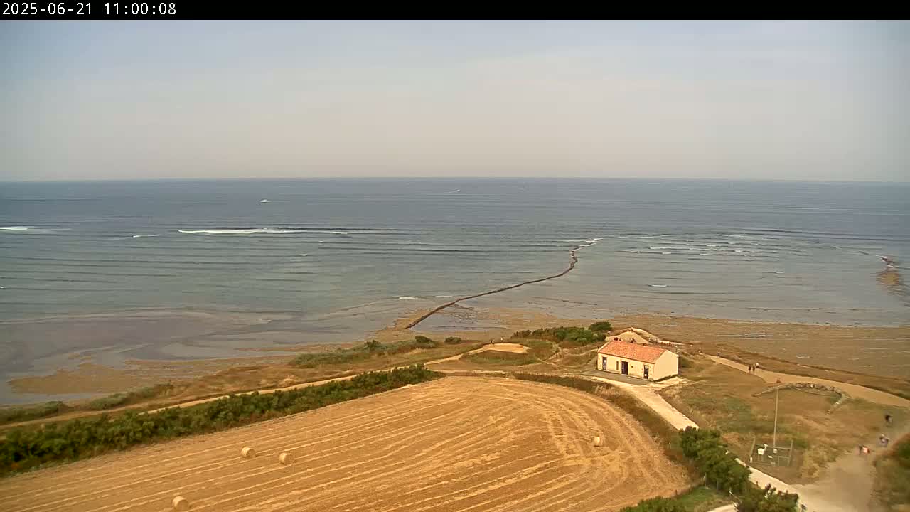 A high-angle view shows a calm ocean with small waves, a small building near a sandy shoreline, and a harvested field with hay bales in the foreground under a hazy sky.