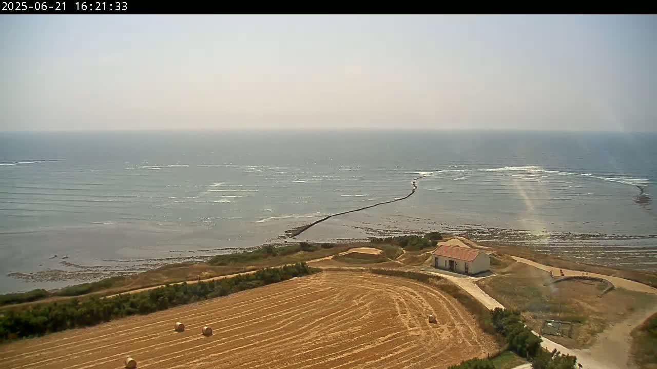 A high-angle view shows a calm ocean under a hazy sky, a small building near the coast, and a harvested field with hay bales.