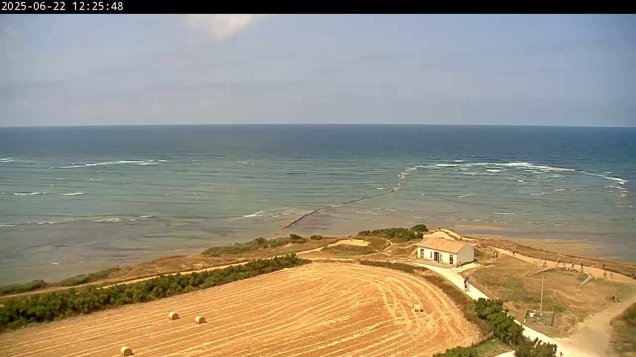A high-angle view shows a coastal scene on a sunny day with a calm sea, a small building near the shore, and a harvested field with hay bales in the foreground.