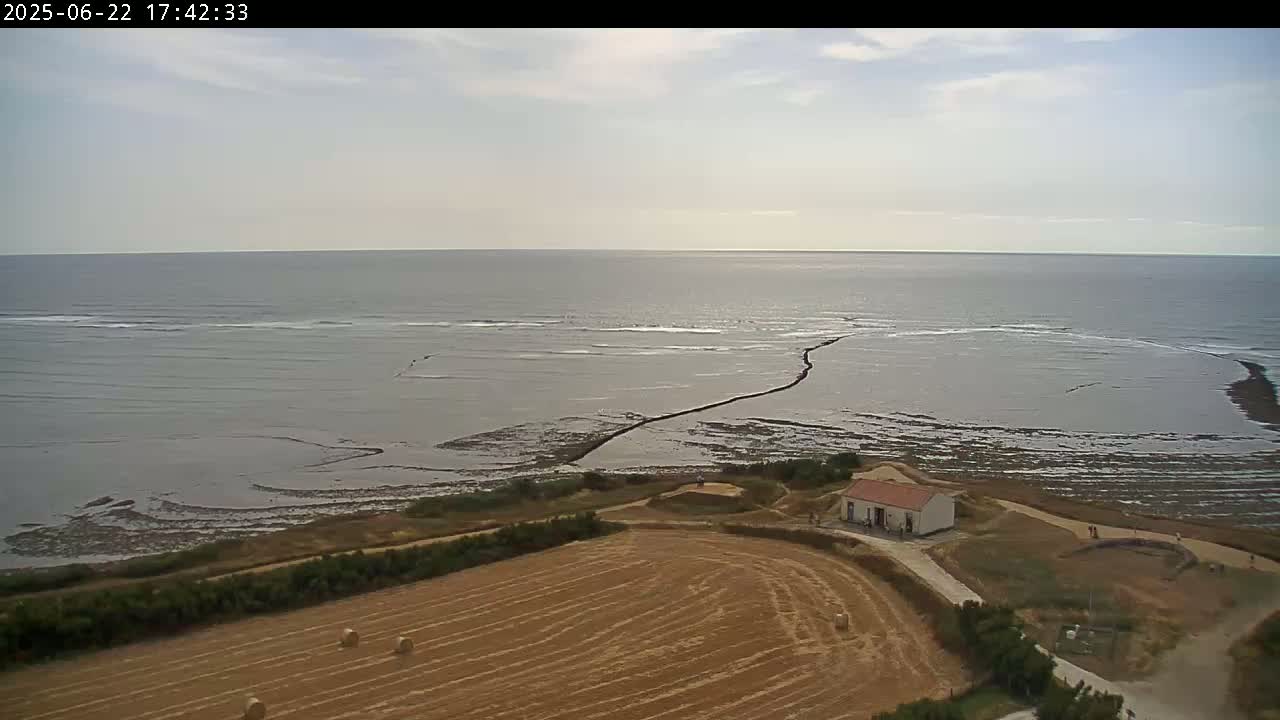 A partly cloudy sky overlooks a calm sea with exposed mudflats near a coastline, where a small building sits beside a harvested field with hay bales.