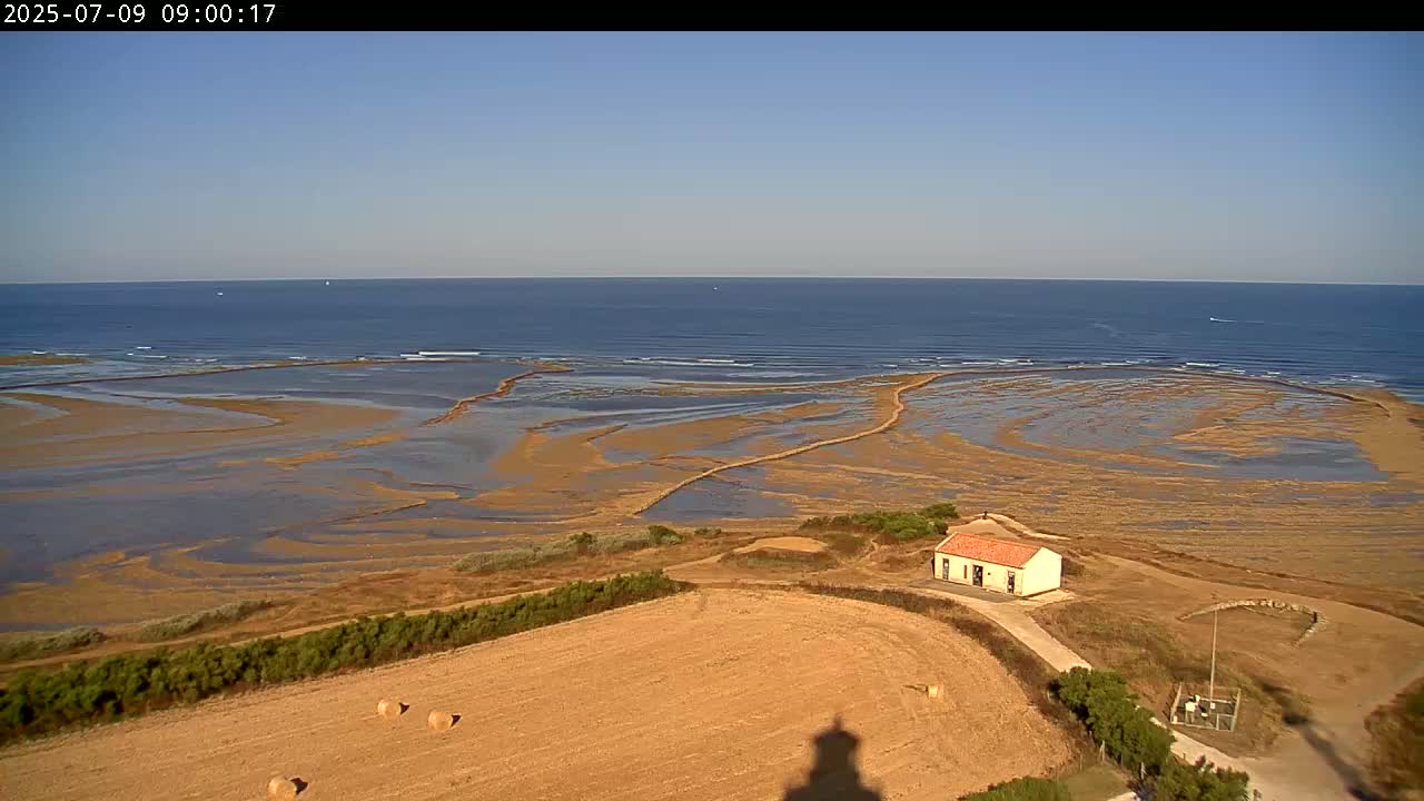 A high-angle view shows a calm ocean under a clear blue sky, with exposed tidal flats and a small building near a field of hay bales.