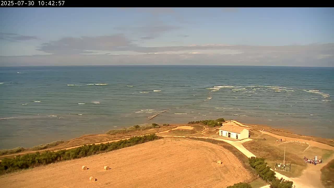 An aerial view shows a small, light-colored building situated on a coastal bluff overlooking a calm sea with small waves under a partly cloudy sky.