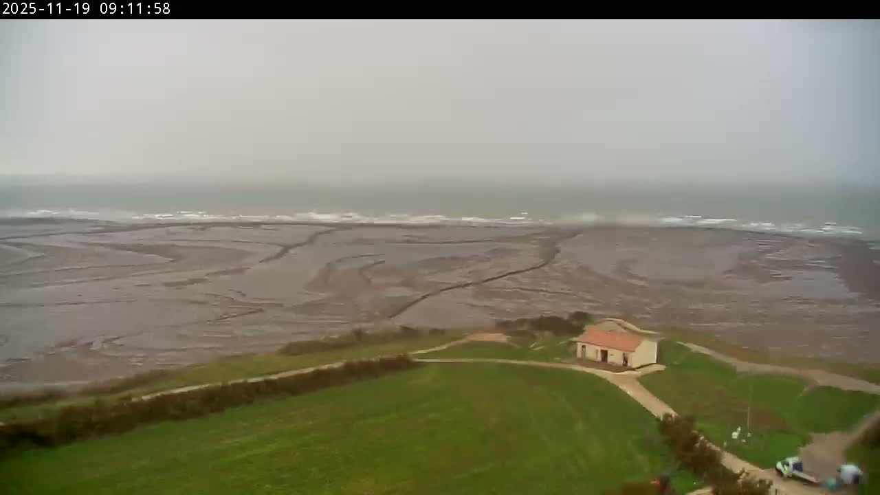 On an overcast day, a wide expanse of muddy tidal flats meets the sea with breaking waves, as seen from a green hillside where a small white house sits beside winding dirt paths.