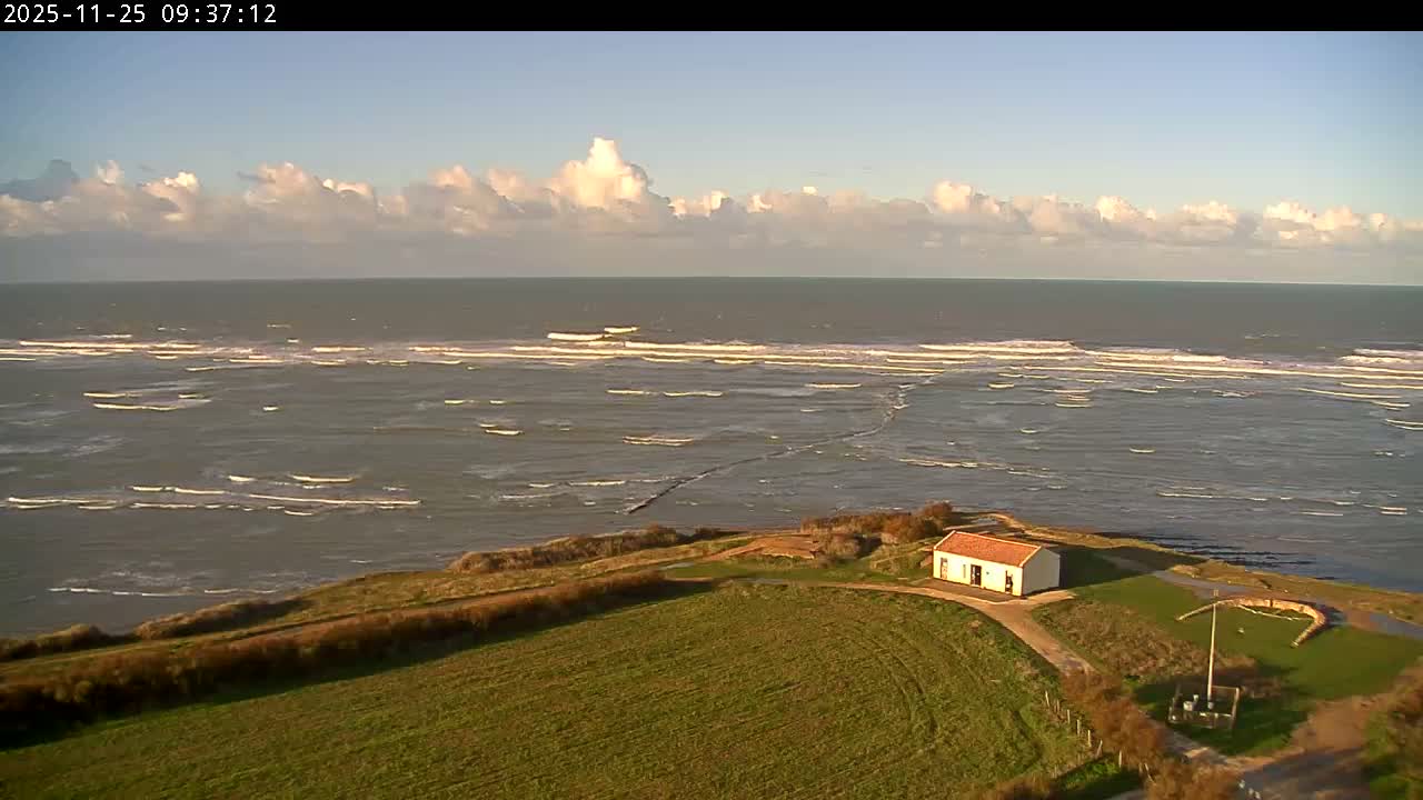 Under a clear and sunny sky with scattered white clouds, a white building with an orange roof sits on a green, grassy coastal bluff overlooking a vast, wavy ocean.
