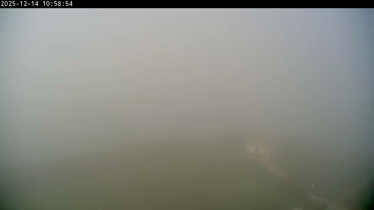 Under a clear and sunny sky with scattered white clouds, a white building with an orange roof sits on a green, grassy coastal bluff overlooking a vast, wavy ocean.