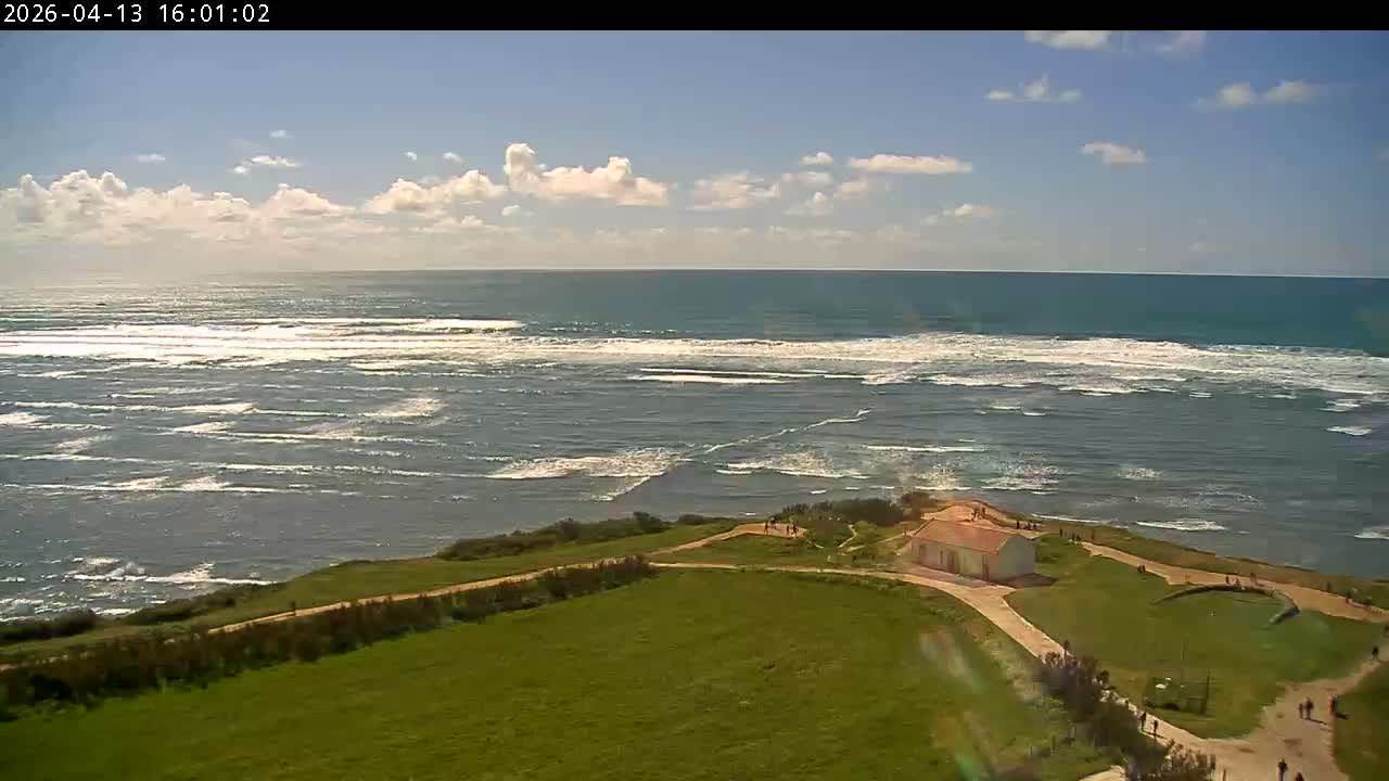 Under a clear and sunny sky with scattered white clouds, a white building with an orange roof sits on a green, grassy coastal bluff overlooking a vast, wavy ocean.