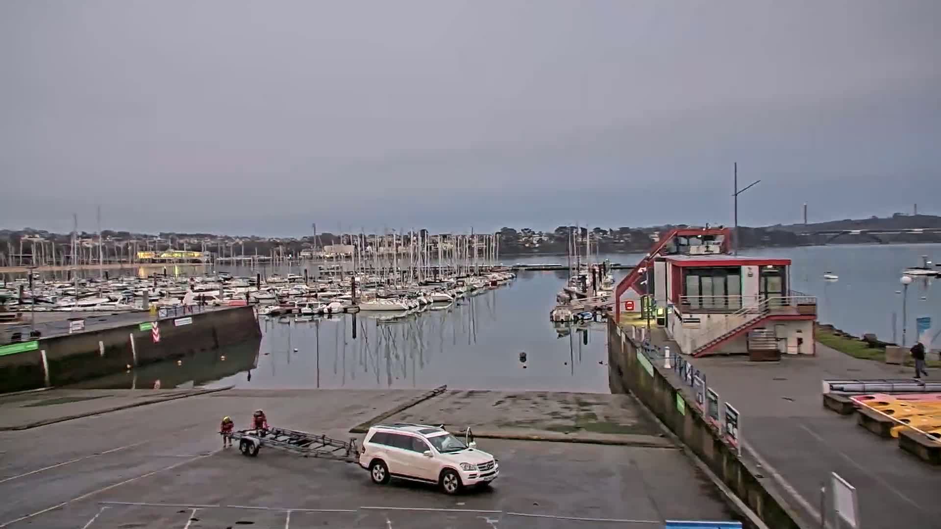 A marina filled with sailboats is viewed from a ramp where a white SUV is being towed by a small trailer, under an overcast sky.