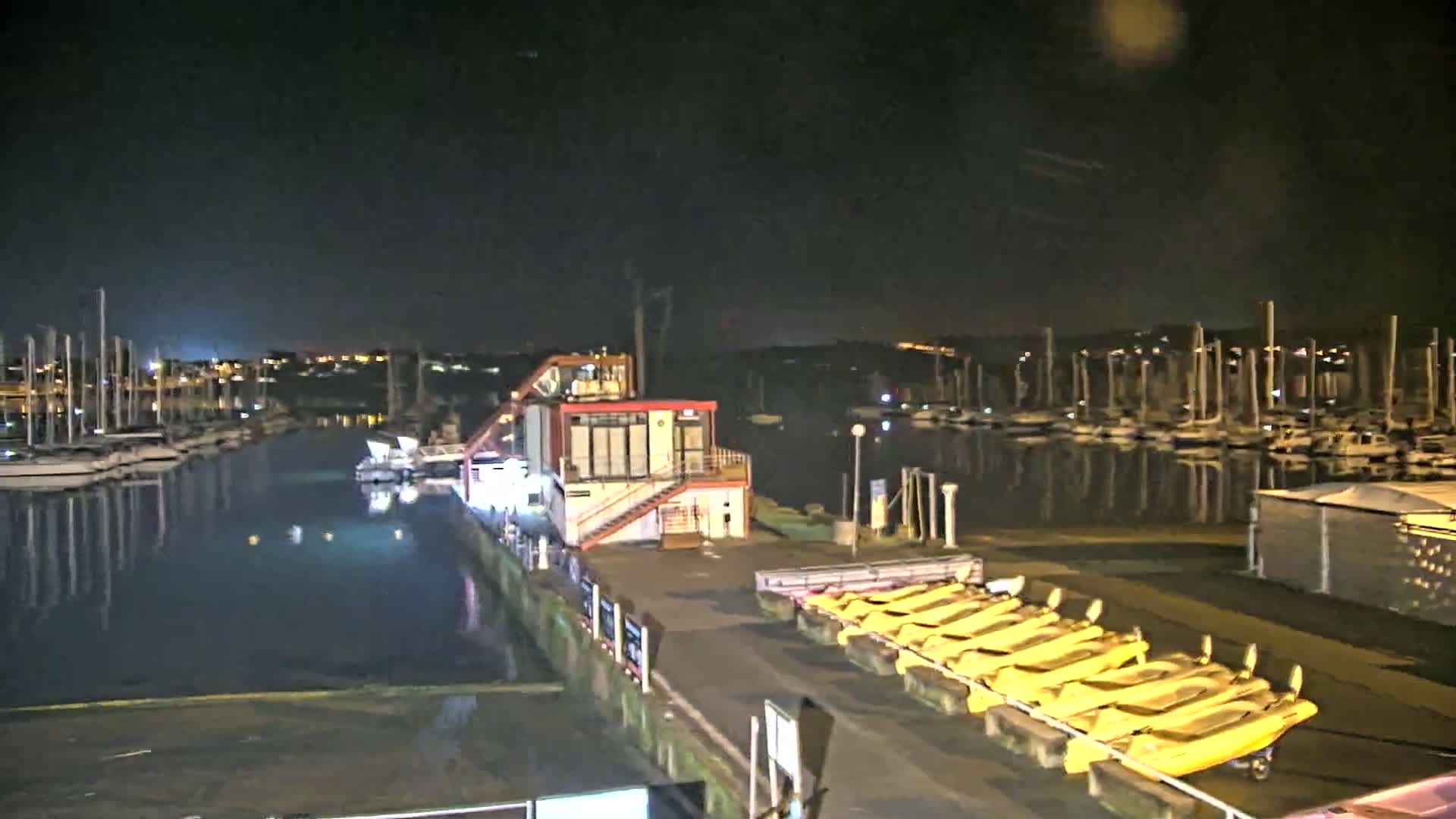 A nighttime view of a marina with numerous docked sailboats, a small building, and a row of yellow boats on the shore under a clear, dark sky.