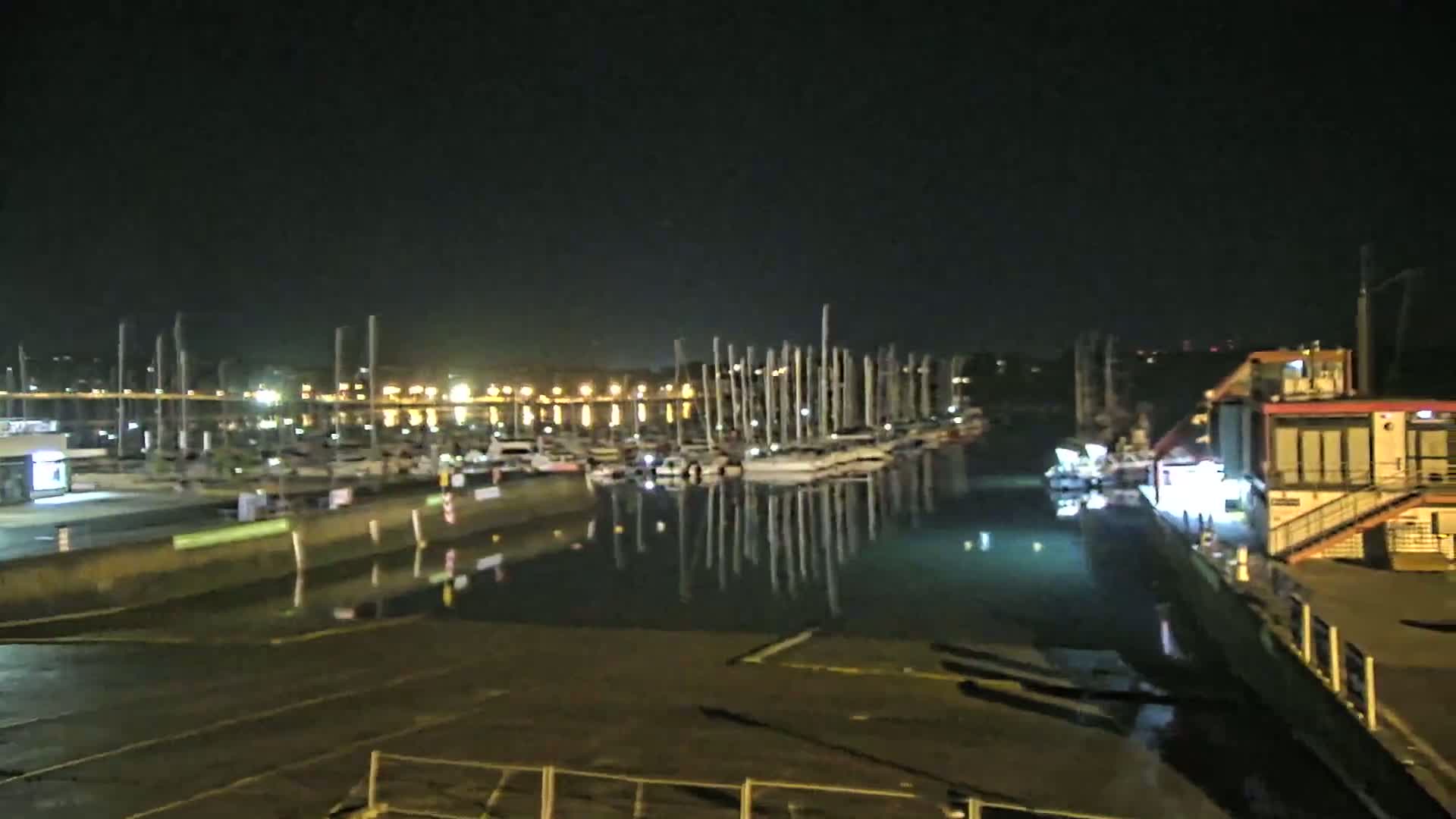 A marina at night, filled with sailboats and illuminated by lights on shore, reflects in the calm water.