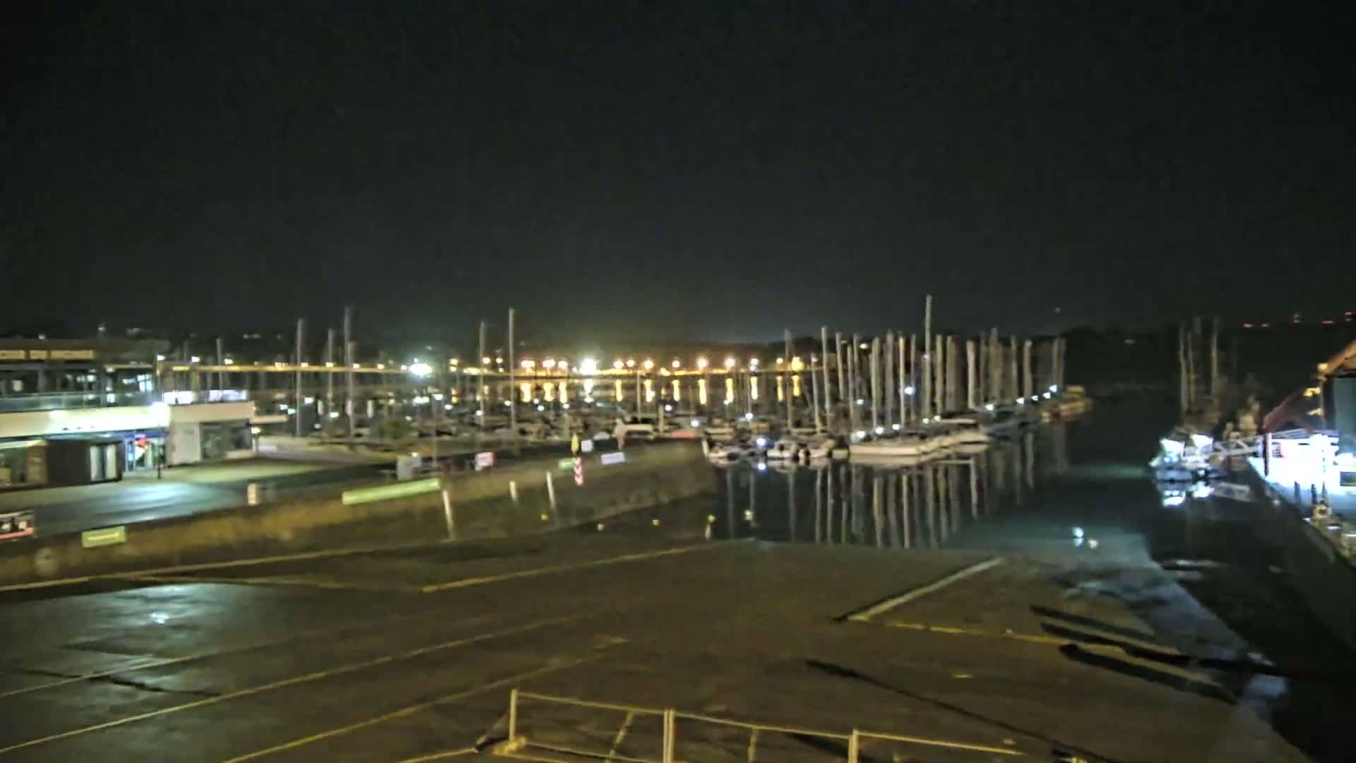 A nighttime view of a marina with numerous sailboats docked, illuminated by lights reflecting on calm water.