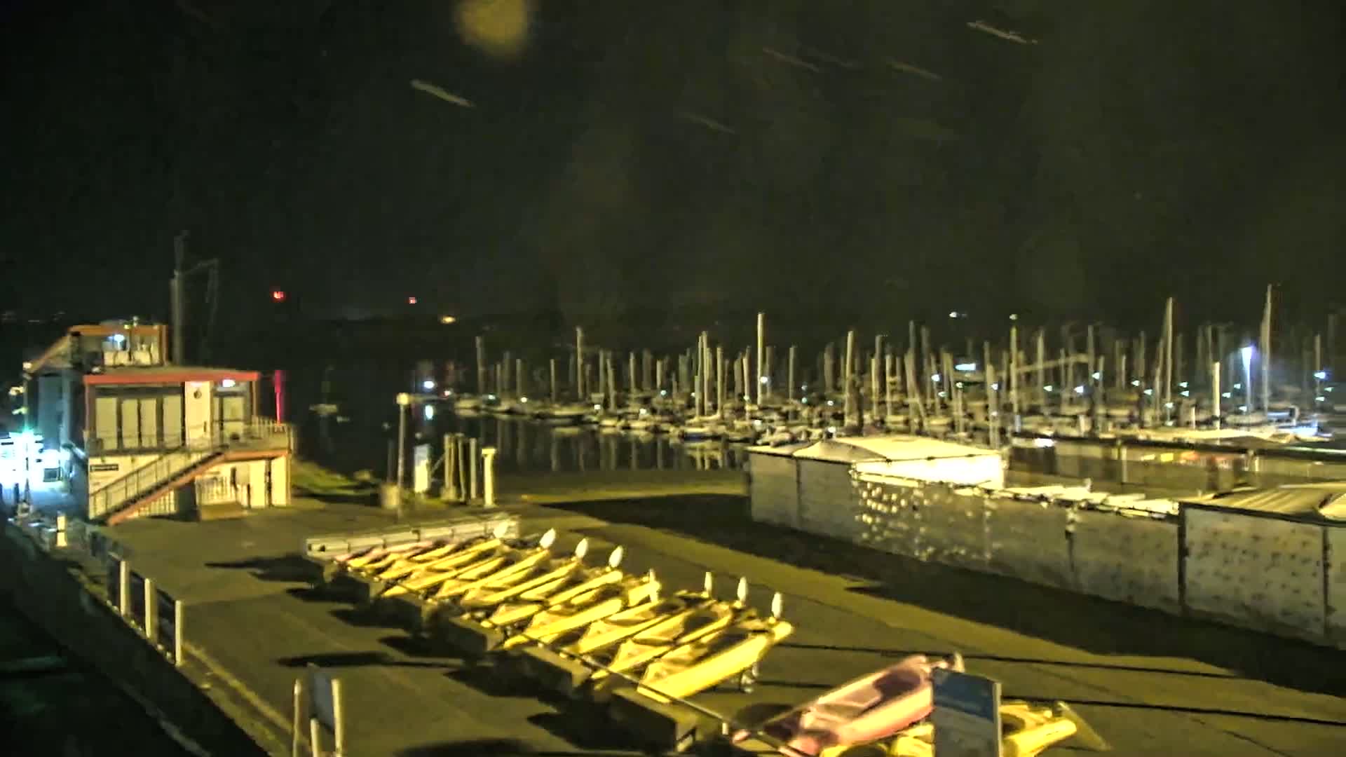 At night, a marina filled with docked sailboats is visible, with numerous yellow kayaks lined up on a nearby dock under a dark, possibly slightly overcast sky.