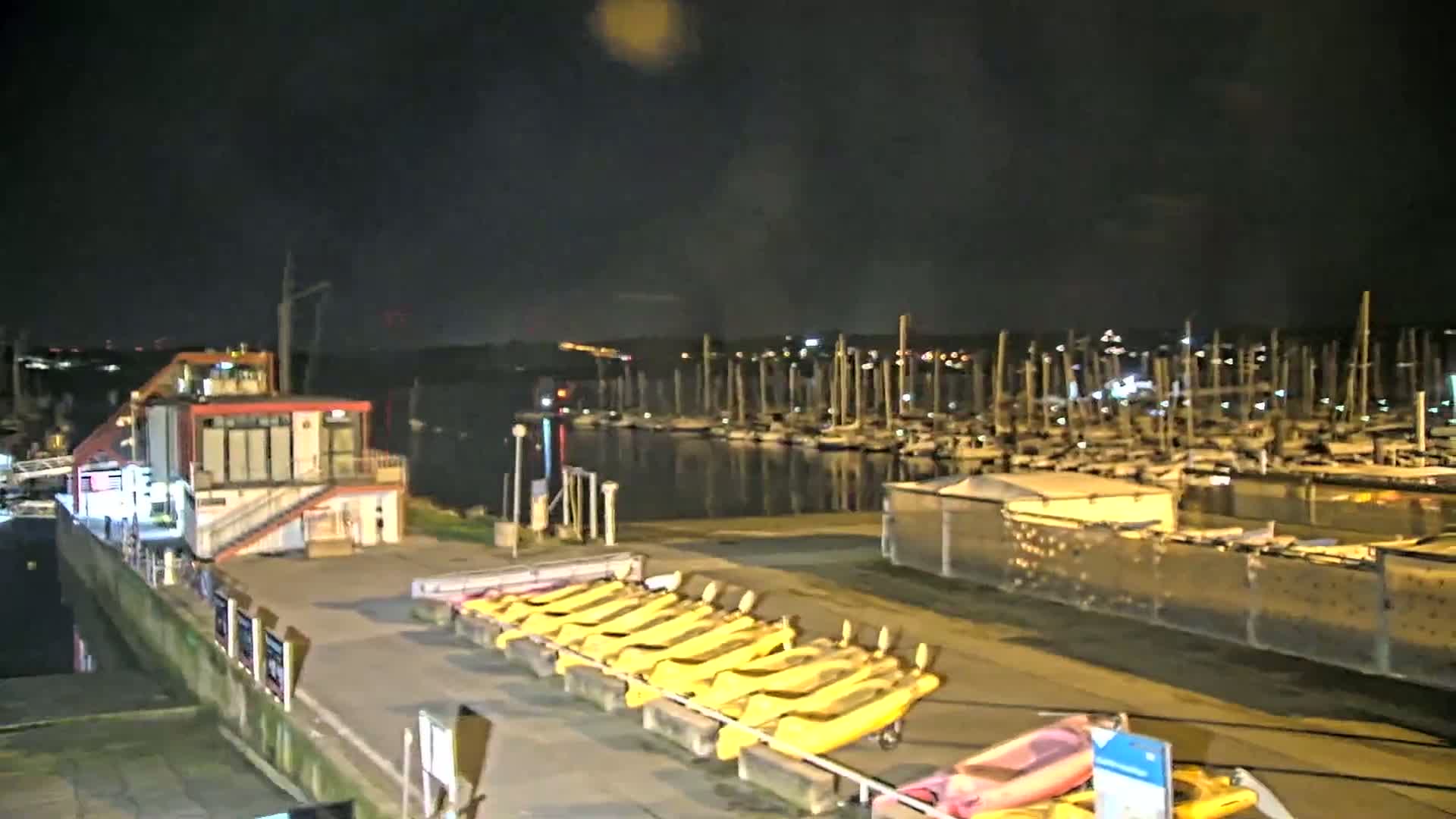 A nighttime view of a marina with numerous boats docked, a small building, and several yellow kayaks lined up on a rack.