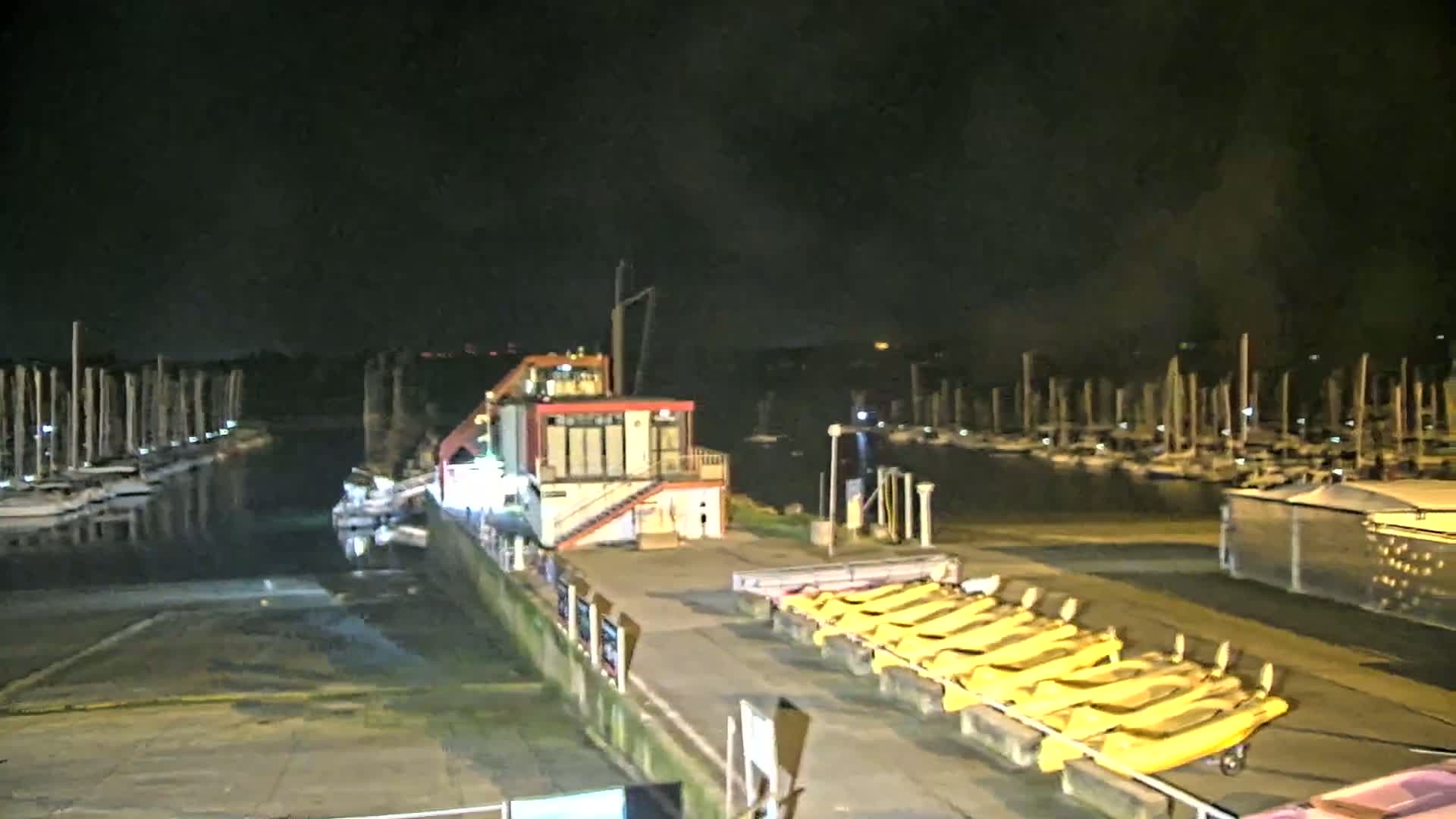 A marina at night, with numerous sailboats docked, a small building, and several yellow boats lined up on a dock.