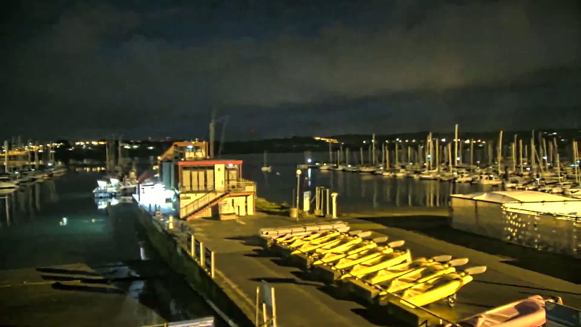 A marina at night, under a dark cloudy sky, shows many boats moored, with a building and several yellow kayaks on a dock.