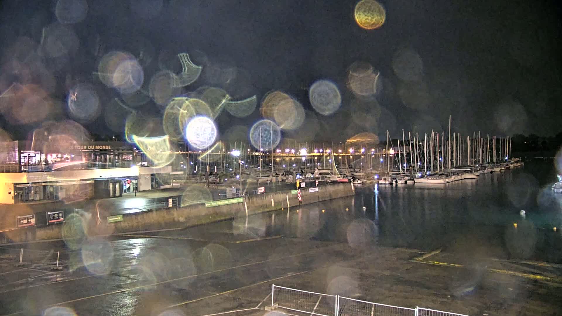 A rain-covered nighttime view of a marina filled with sailboats, with a building and parking lot in the foreground.