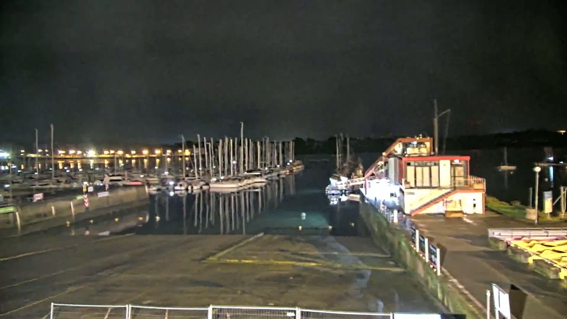 At night, numerous sailboats are docked in a marina, with a building and a paved area visible nearby.