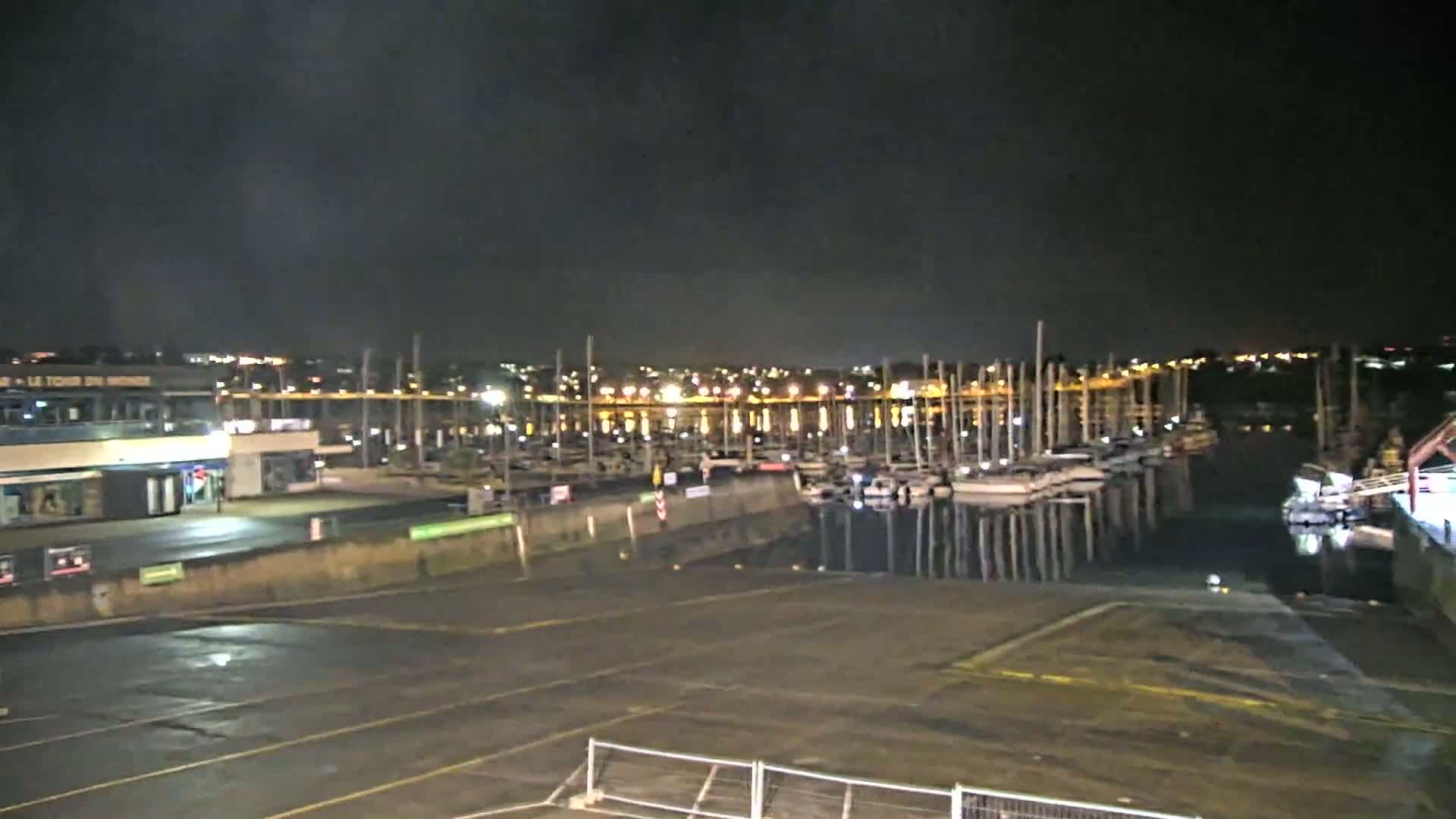 At night, a marina filled with sailboats is visible across a dark, empty parking lot under a cloudy sky.