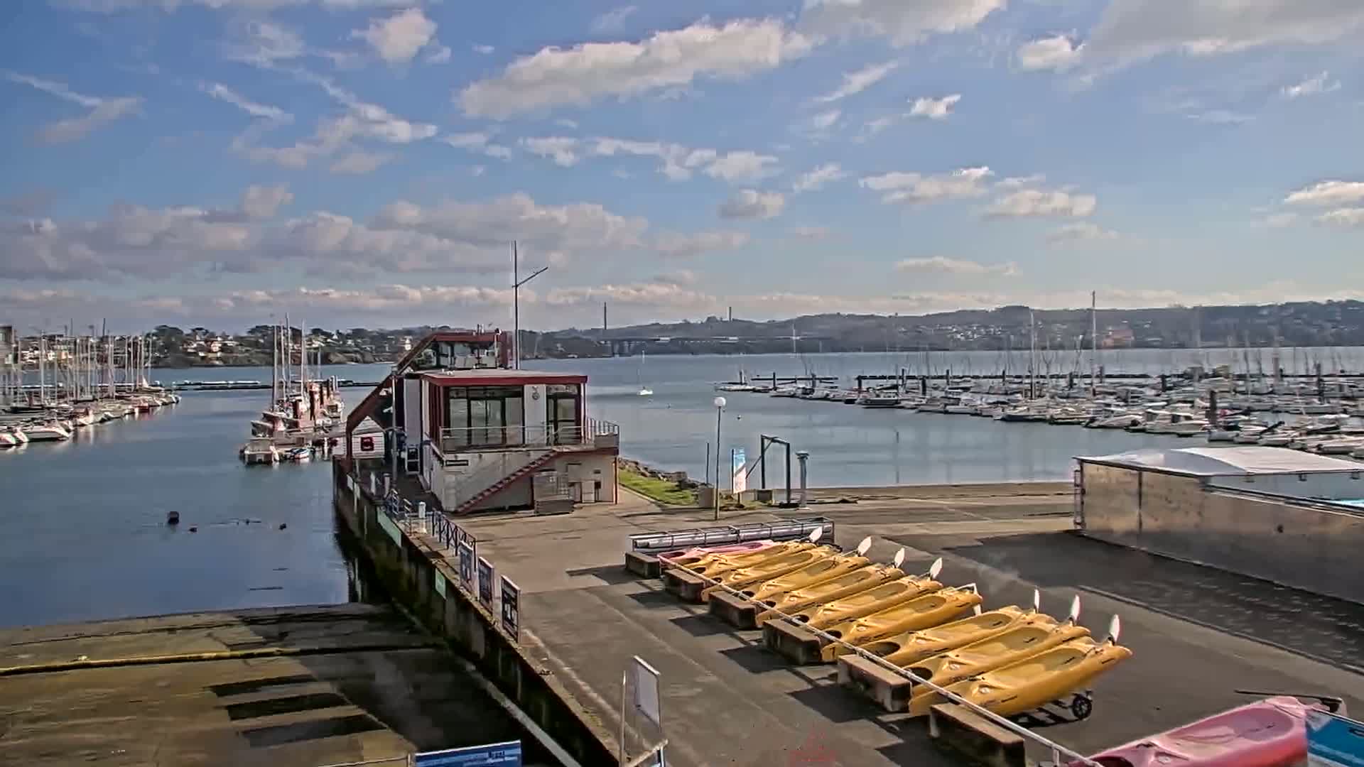 A marina with numerous boats, a small building, and rows of yellow kayaks is shown under a partly cloudy sky.