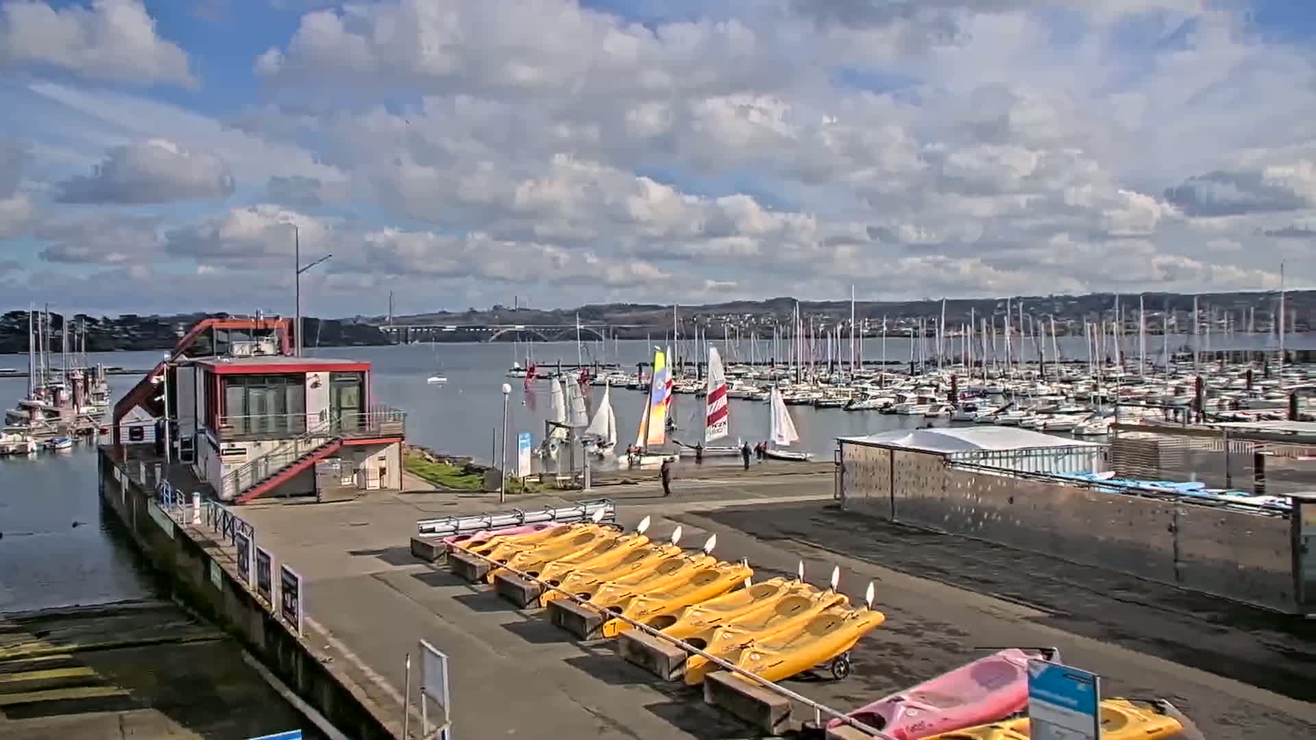 A marina with numerous sailboats and kayaks is shown on a partly sunny day with some clouds.