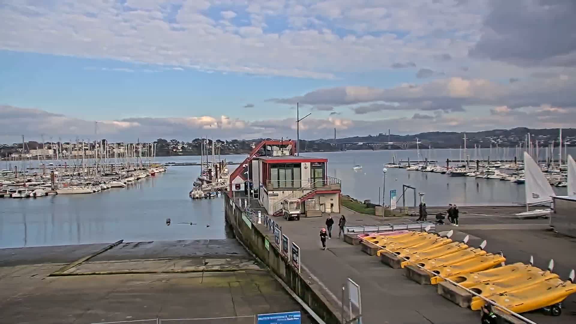 A marina filled with sailboats under a partly cloudy sky, features a small red building with a walkway leading to several yellow kayaks.