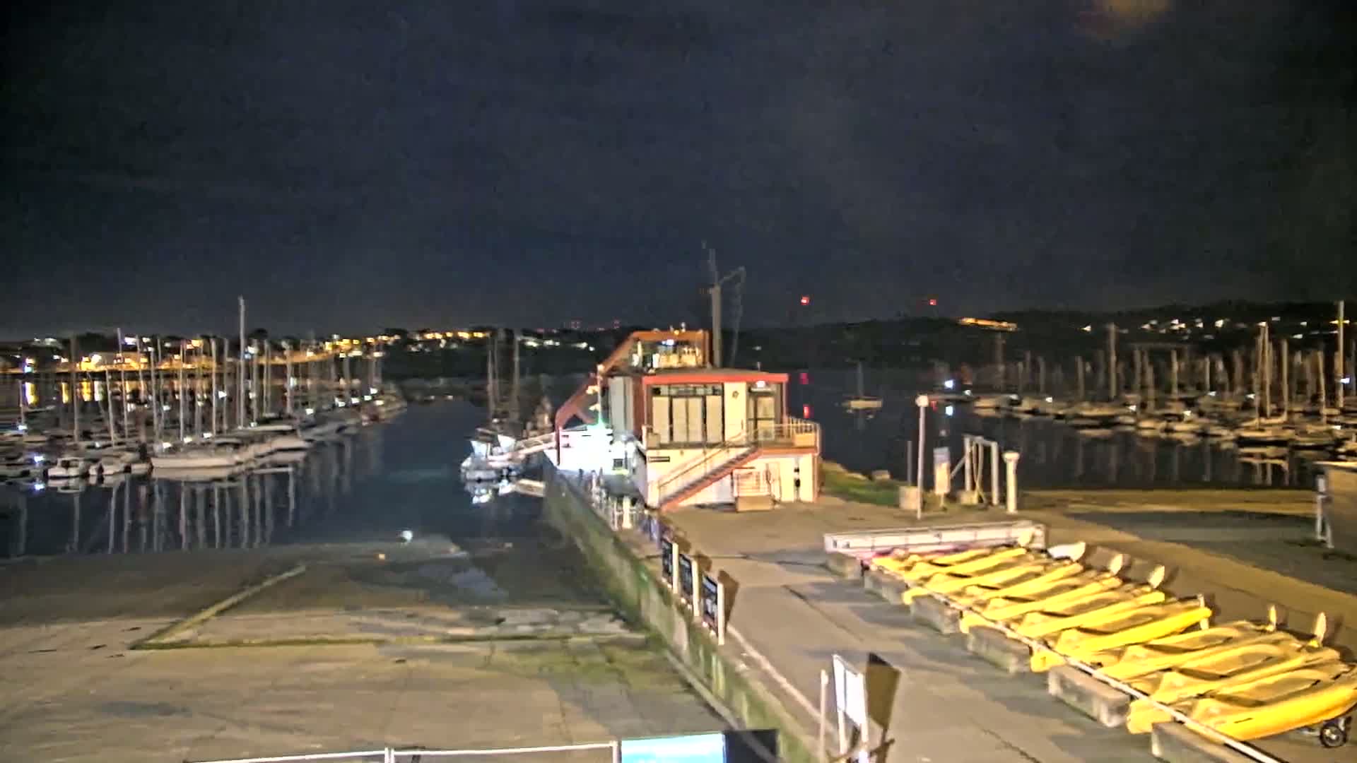 A marina at night, under a dark sky, shows many sailboats docked and a building with yellow boats lined up nearby.