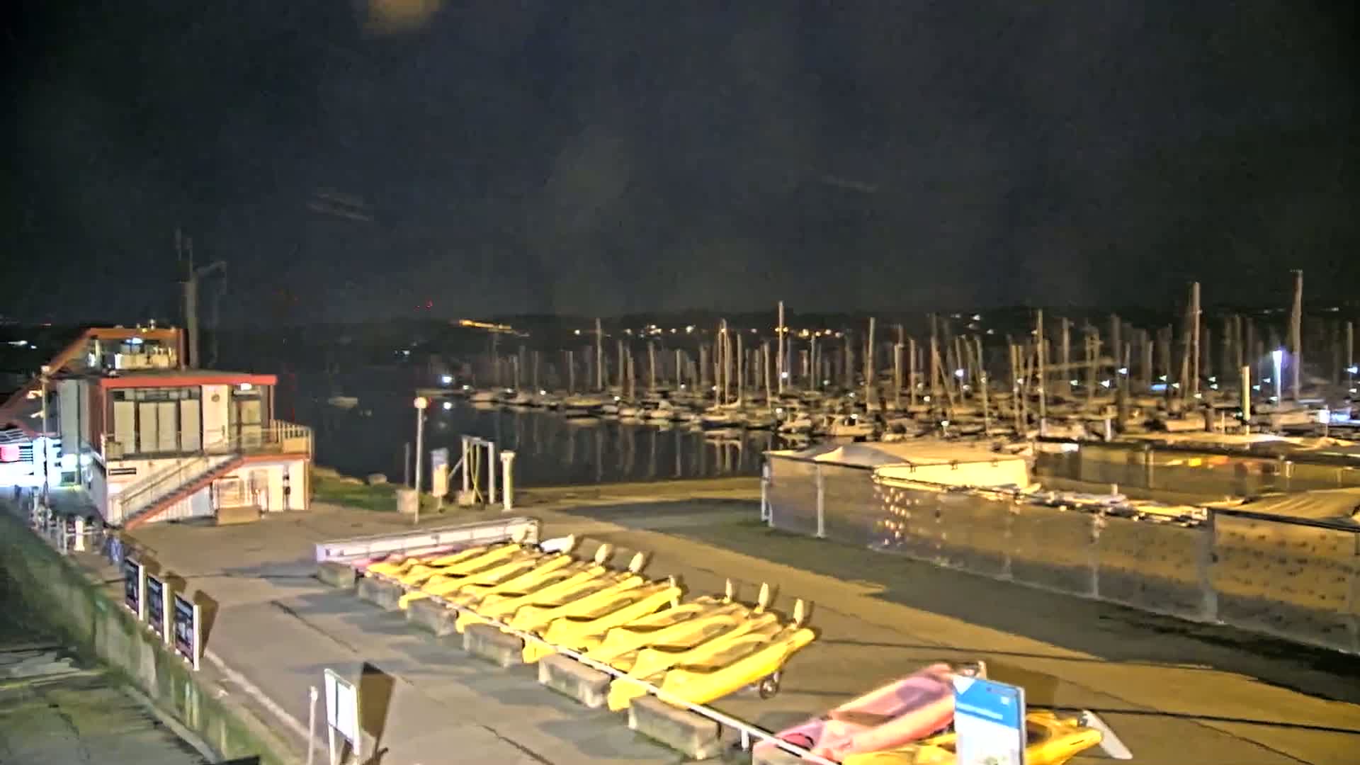 A nighttime view of a marina with many boats docked, a small building, and several yellow kayaks lined up on a rack.