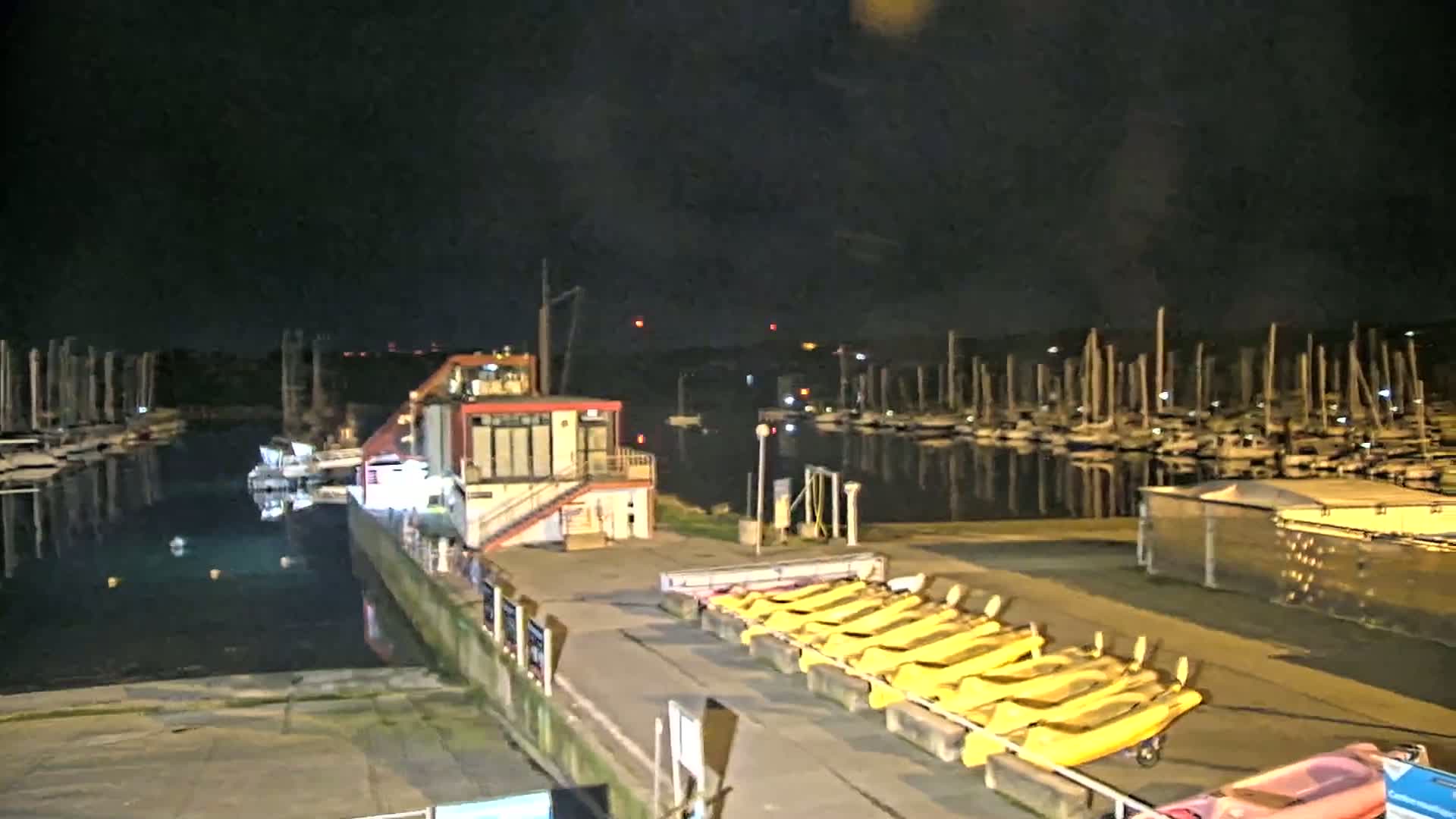 A nighttime view of a marina with numerous boats docked, a small building, and several yellow kayaks lined up on a paved area.