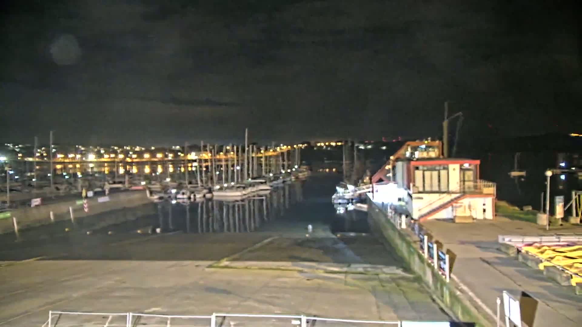 A nighttime view of a marina with numerous boats docked, a small building, and a city skyline illuminated in the background under a dark, cloudy sky.