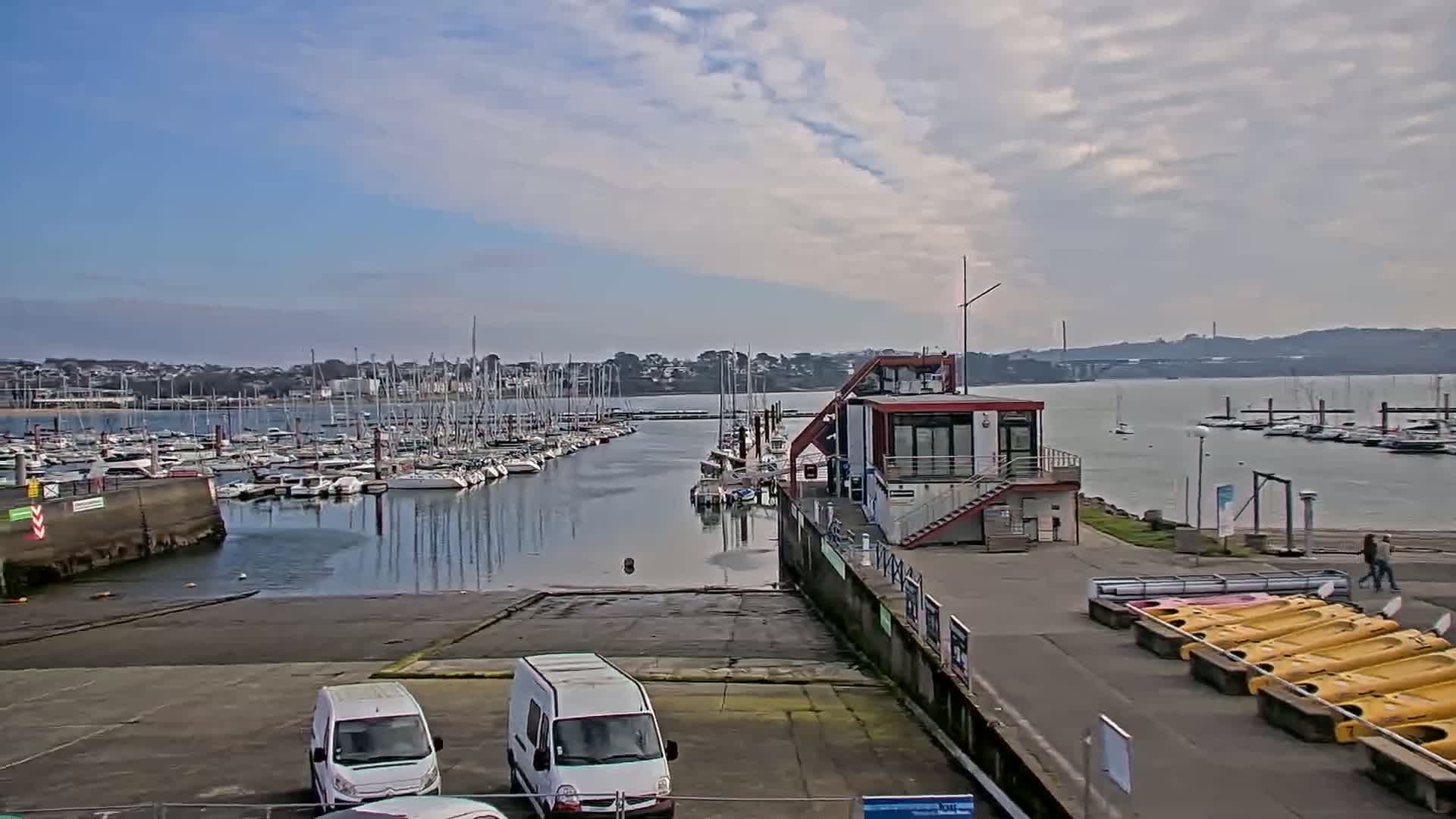 A marina filled with sailboats under a partly cloudy sky, with a small building and several vans parked nearby.
