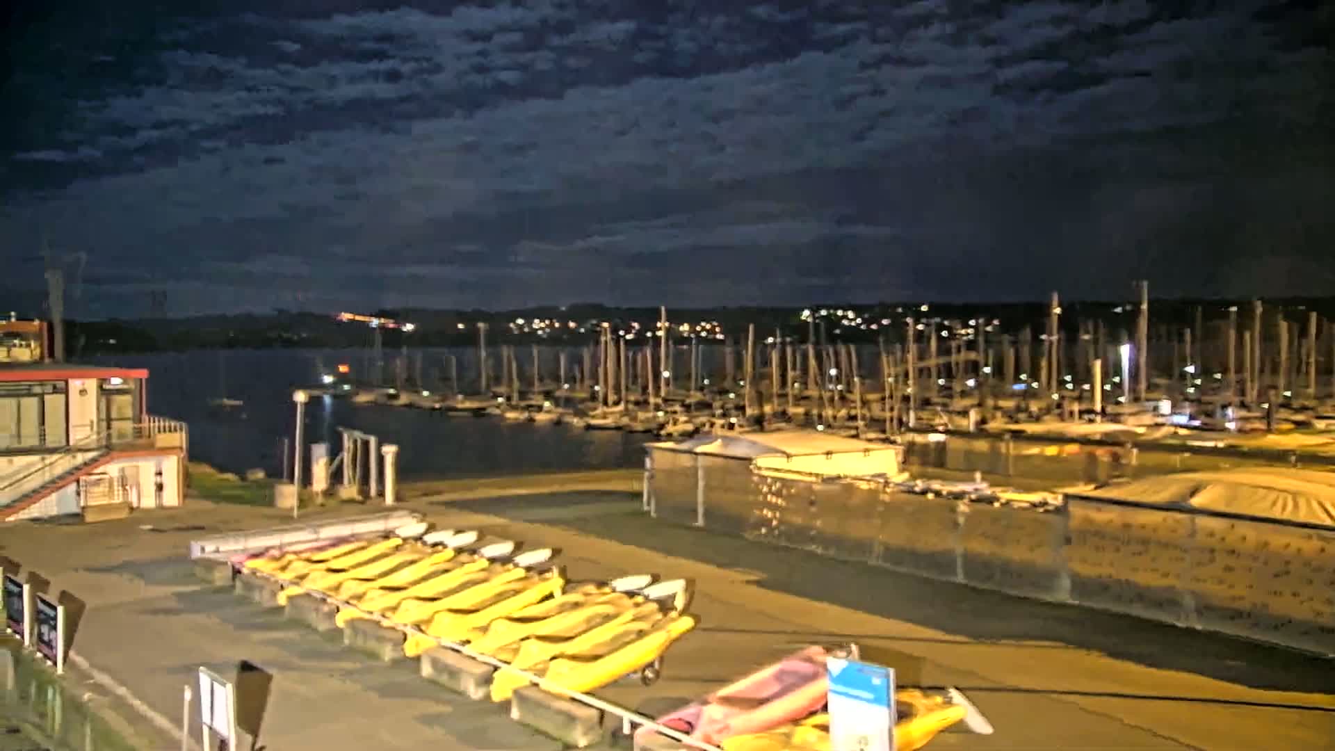 Under a partly cloudy night sky, a marina filled with numerous sailboats is visible across a body of water from a dock where yellow kayaks are stored.