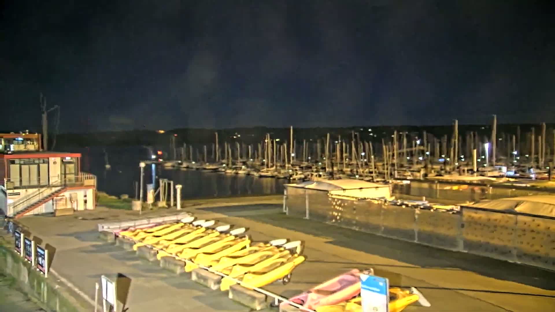At night, a marina filled with docked sailboats is visible across a body of water, with a row of kayaks lined up on the shore nearby, under a dark sky.