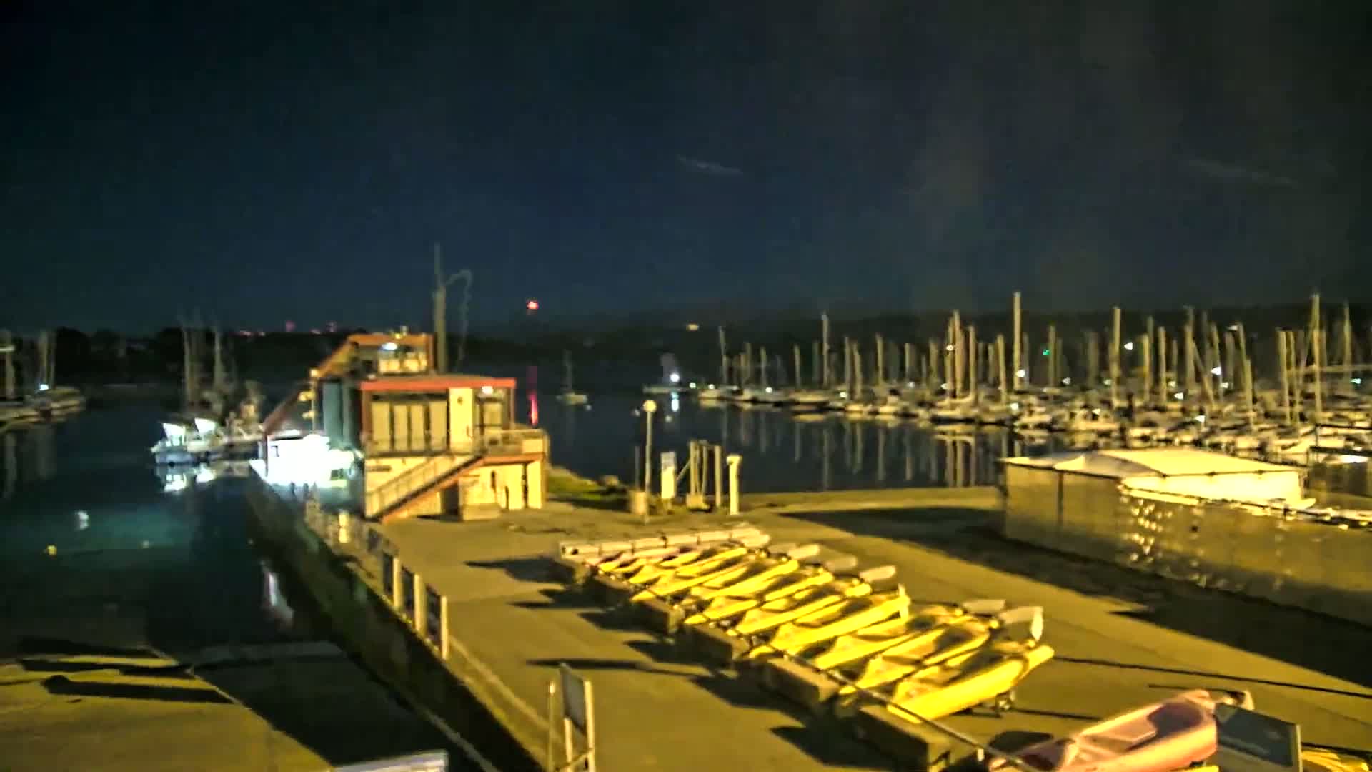 A marina at night, illuminated under a dark sky, shows numerous boats moored in the water and several yellow kayaks lined up on a dock.