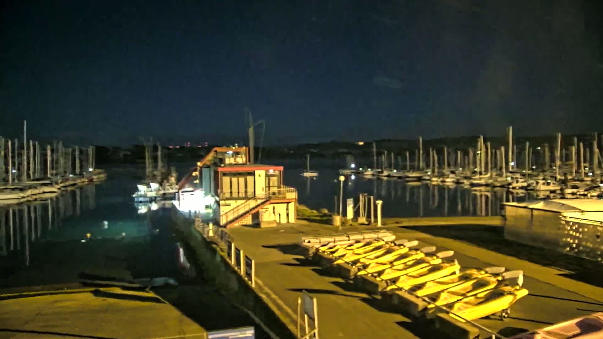 At night, a marina with numerous boats docked reflects in calm water, illuminated by artificial lights, with several yellow kayaks lined up on a nearby dock next to a small building.
