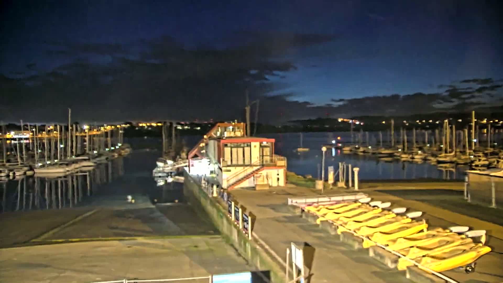 A marina at night under a partly cloudy sky shows many sailboats docked, a small building with lights on, and a row of yellow small boats on the shore.