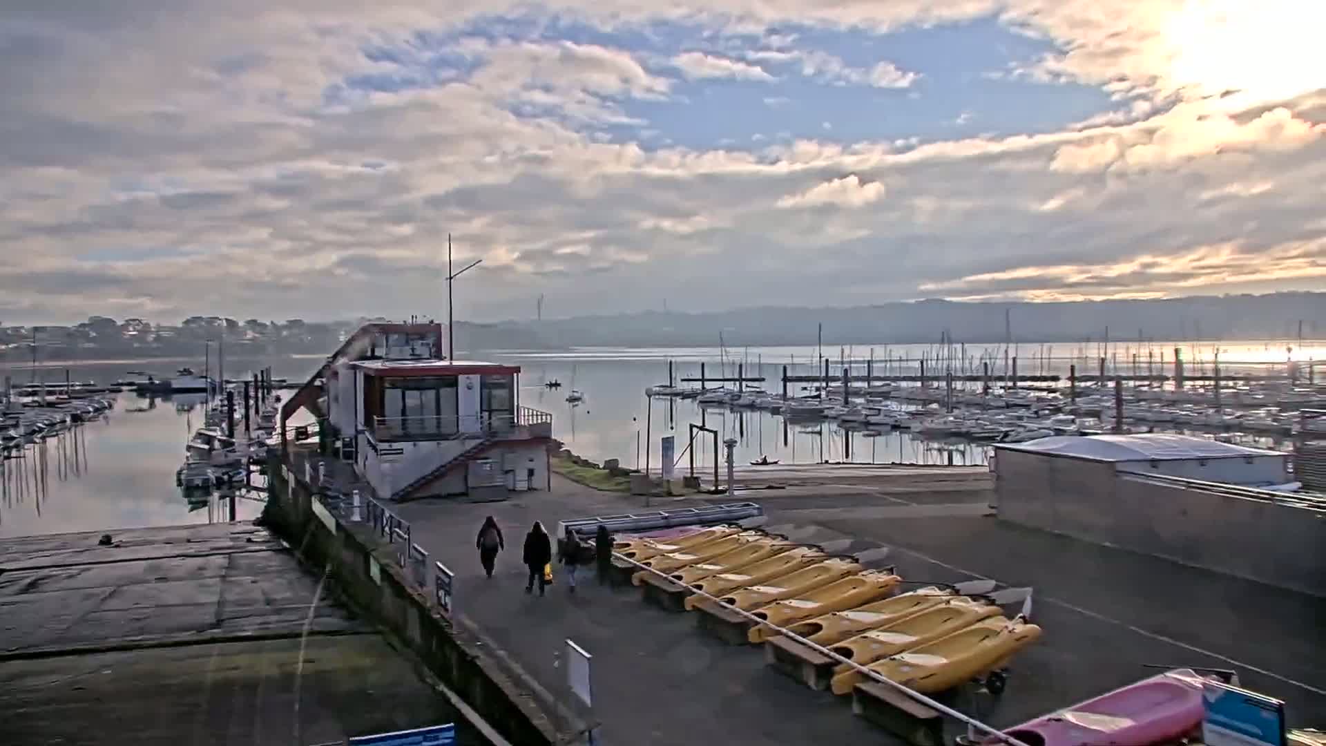 A marina filled with sailboats under a partly cloudy sky, with a small building and several kayaks lined up on the dock.