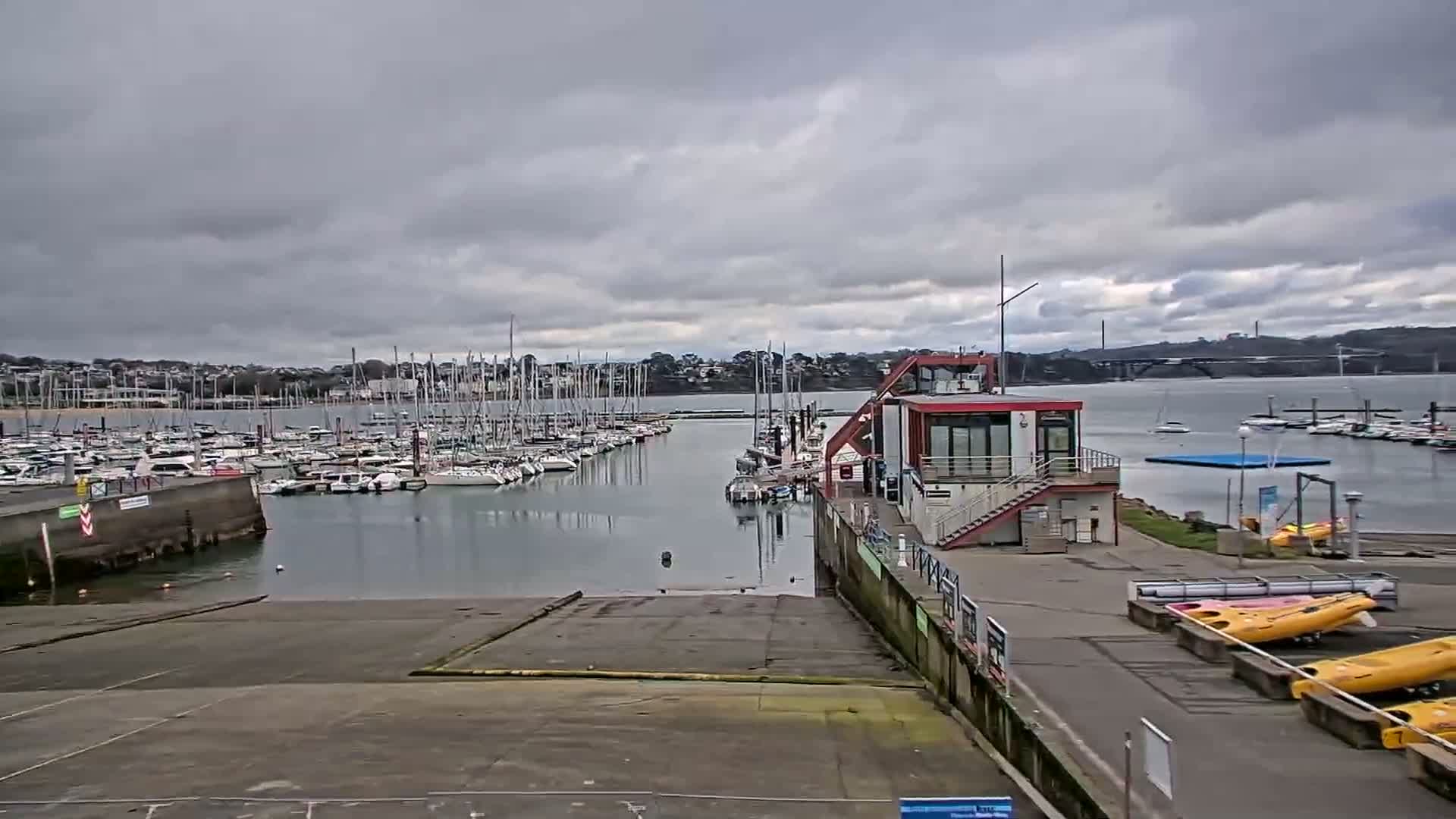 A marina filled with sailboats under a cloudy sky, with a modern building and several kayaks visible on the shore.