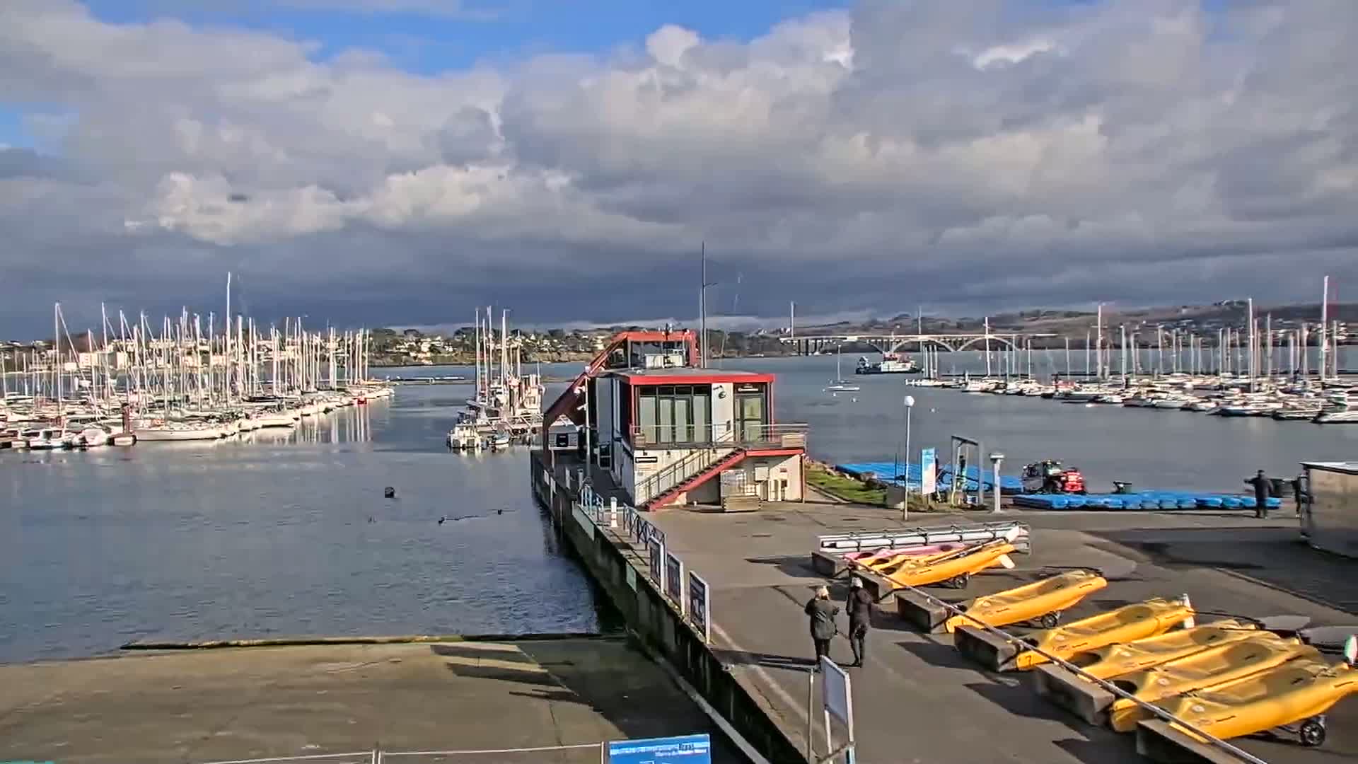 A marina filled with sailboats under a partly cloudy sky, featuring a small building with kayaks lined up on the dock.