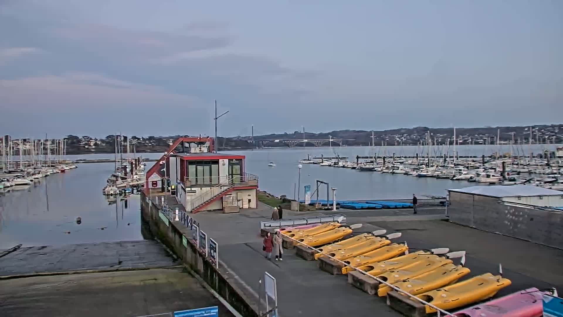 A marina with numerous sailboats and a building with kayaks in the foreground under an overcast sky.