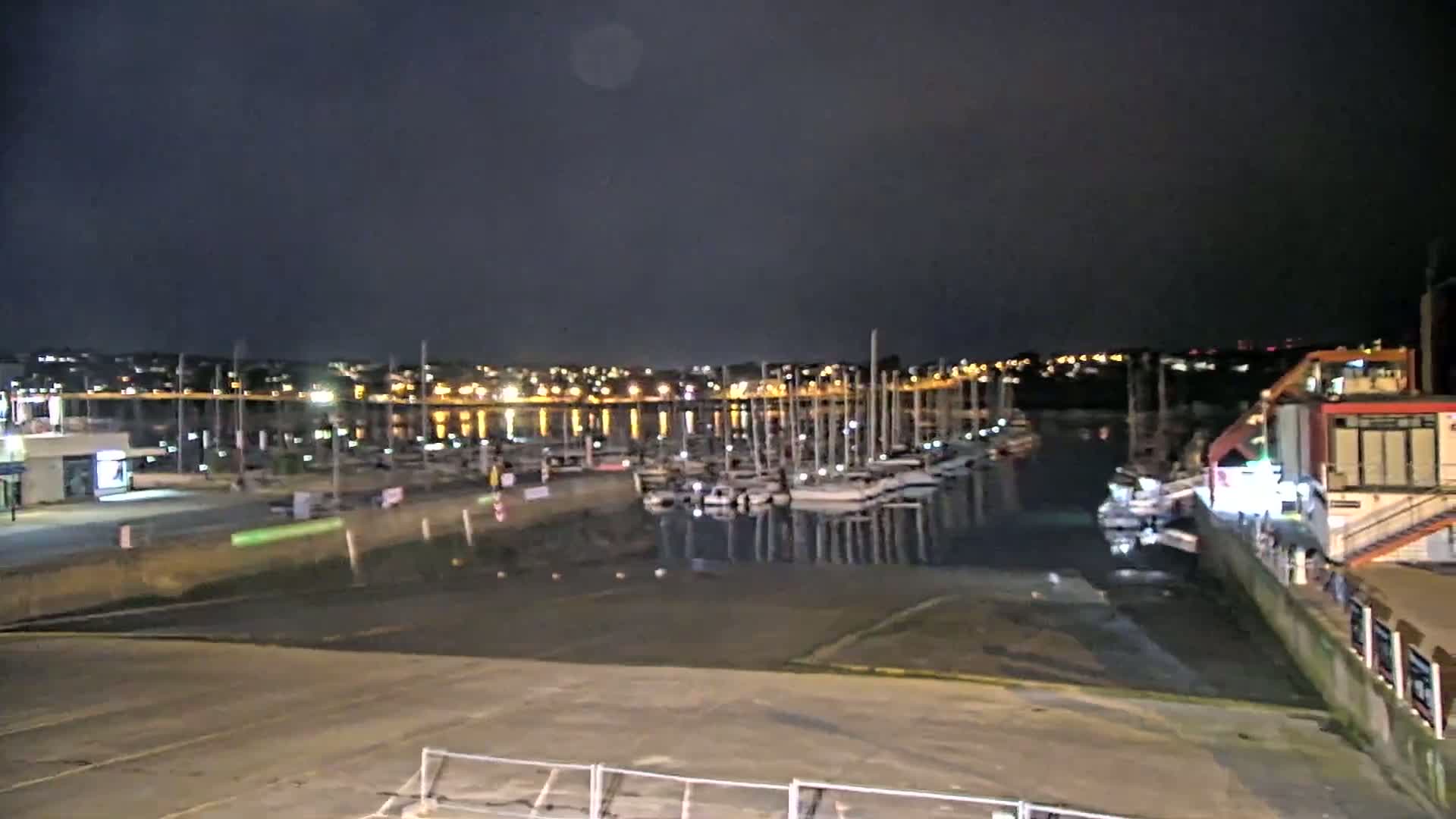A nighttime view of a marina with numerous sailboats docked, under a dark, cloudy sky.