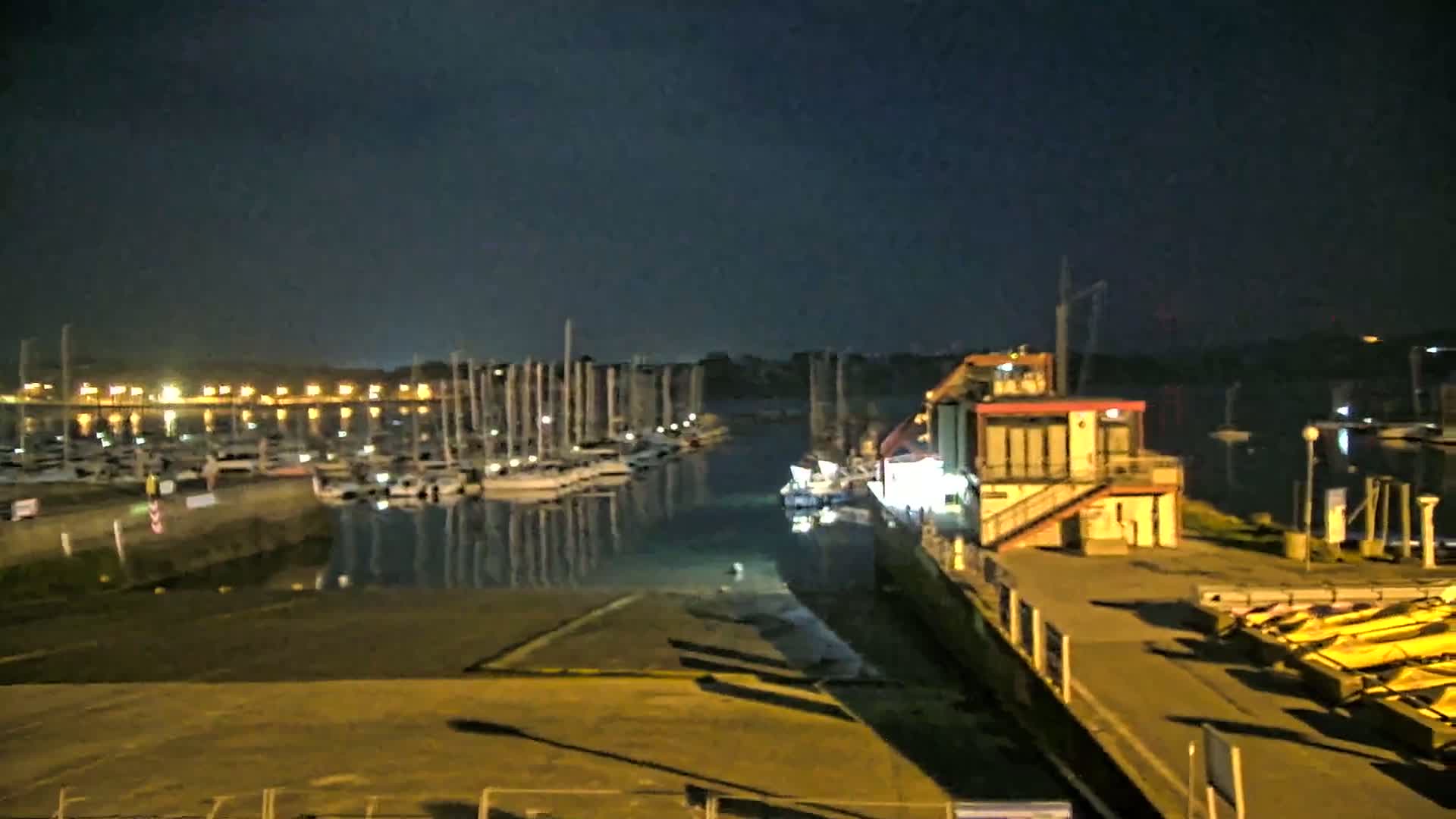 A marina at night under a dark sky, with numerous boats docked and lights illuminating the waterfront.