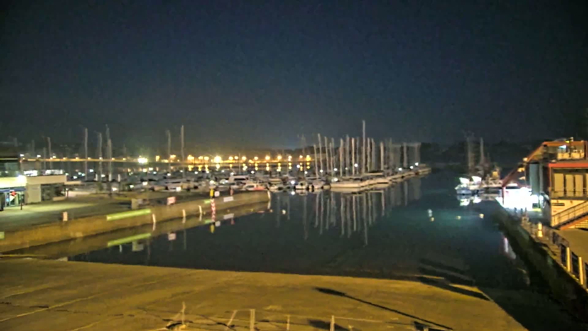 A nighttime view of a marina with numerous boats docked, their masts reflecting in the calm water under a dark sky.