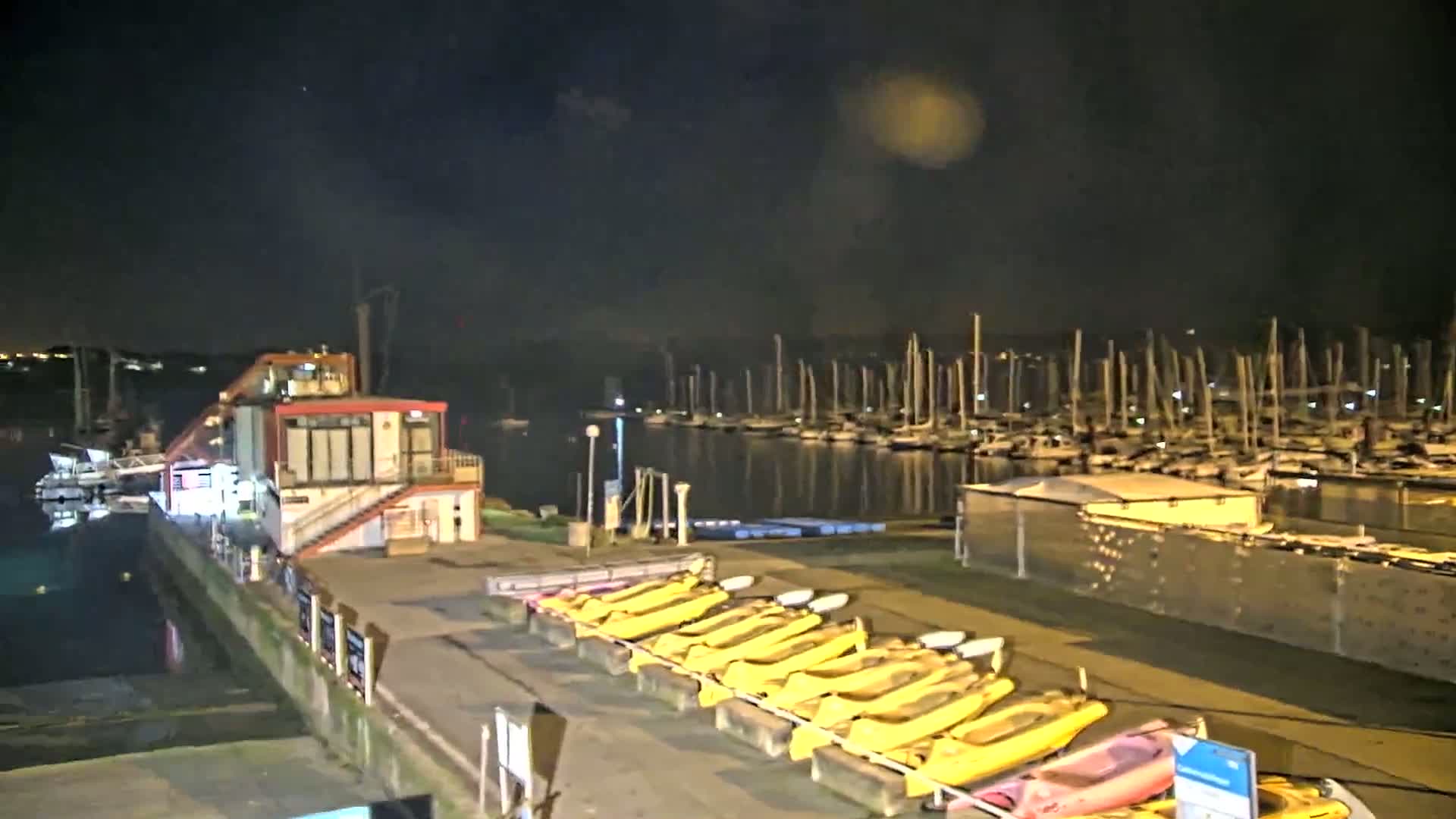 A marina at night, under a dark sky, shows many sailboats moored in the water, with kayaks lined up on the dock next to a small building.