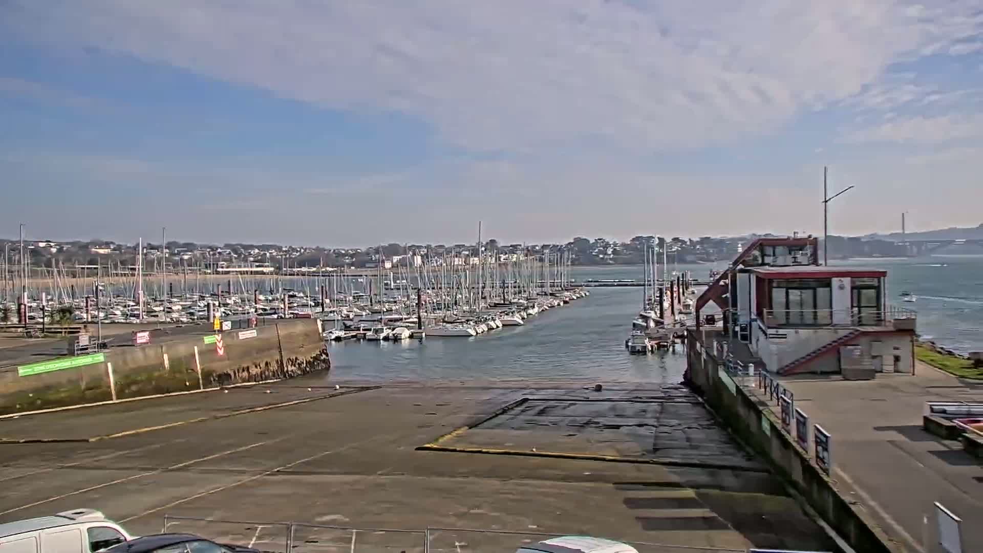 A marina filled with sailboats is seen on a partly cloudy day, with a small building and paved area in the foreground.