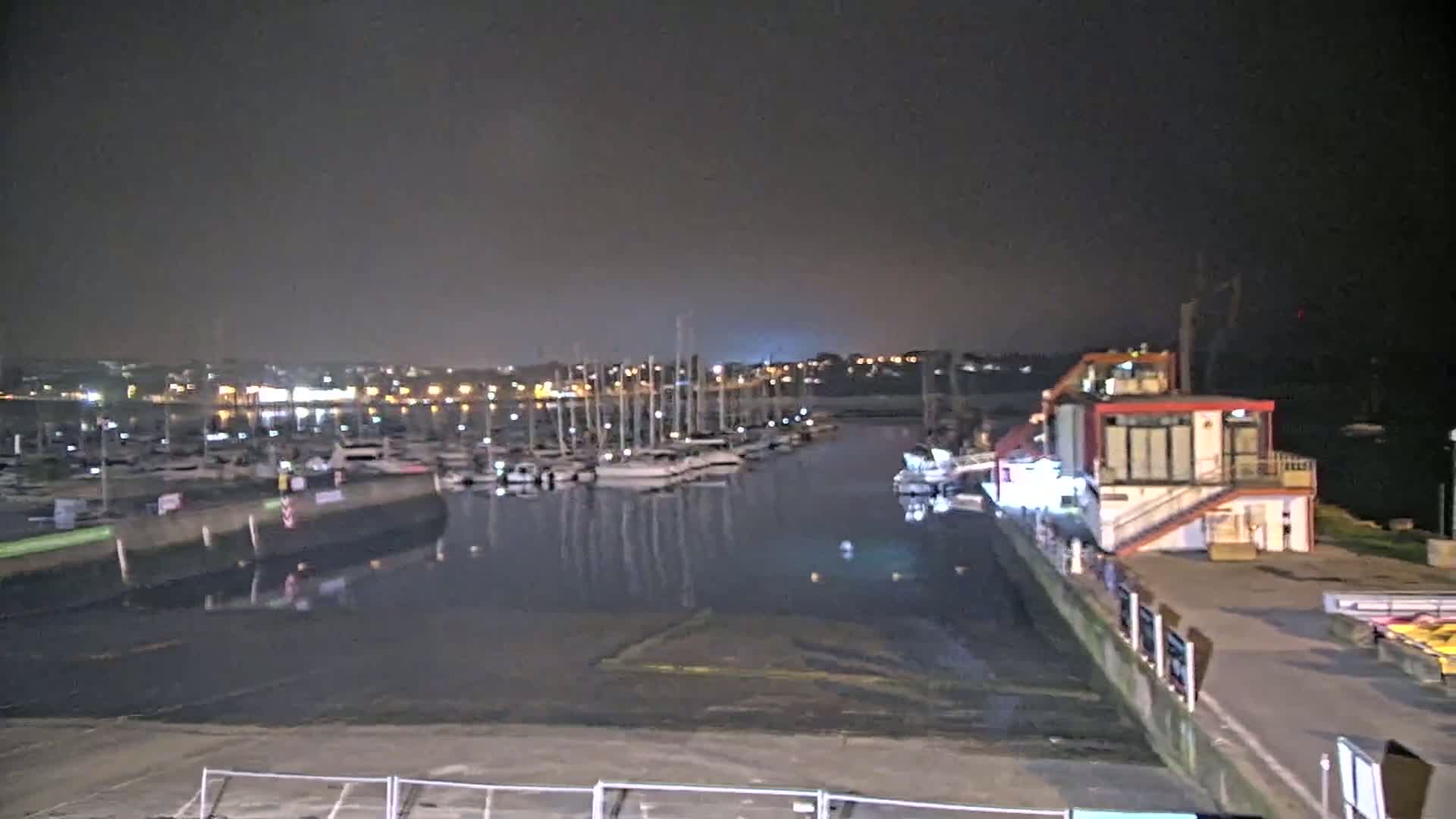 A marina at night, filled with many docked sailboats, is lit by artificial lights under a dark sky.