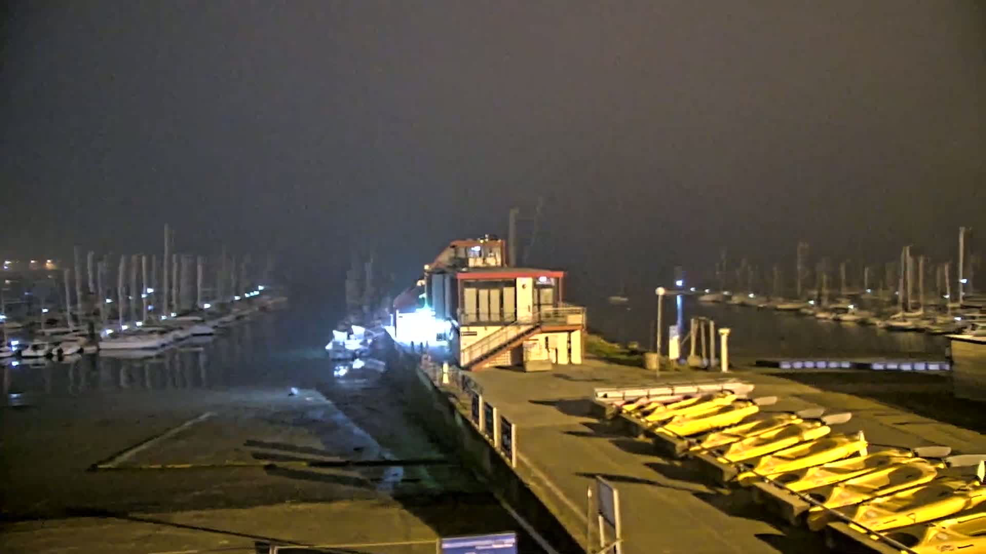 A foggy night scene shows a marina with numerous boats moored, a building with lights on, and a row of yellow kayaks lined up on a dock.