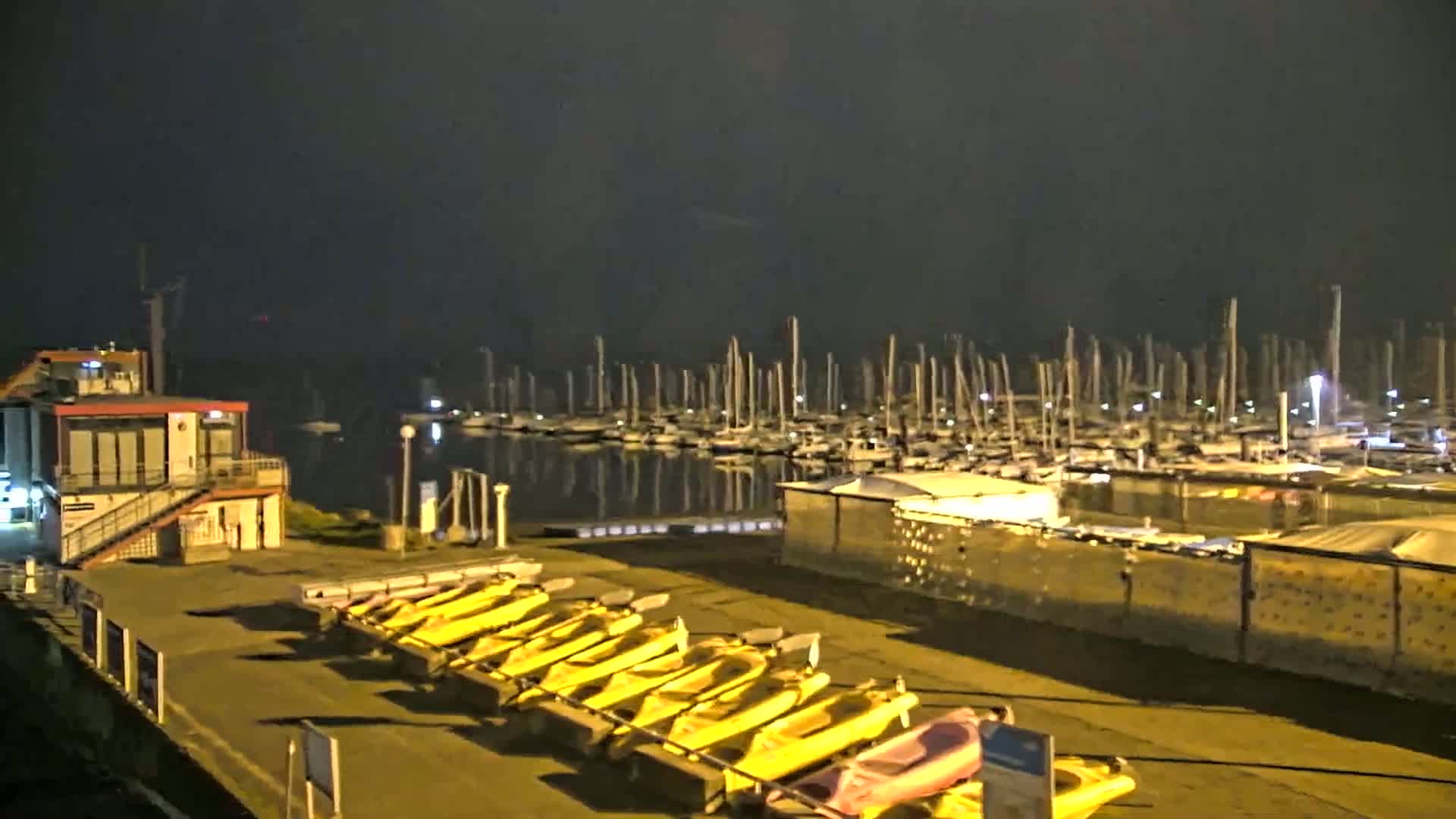 At night, under a dark sky, a marina filled with docked sailboats is visible across the water from a small building and a row of yellow kayaks.